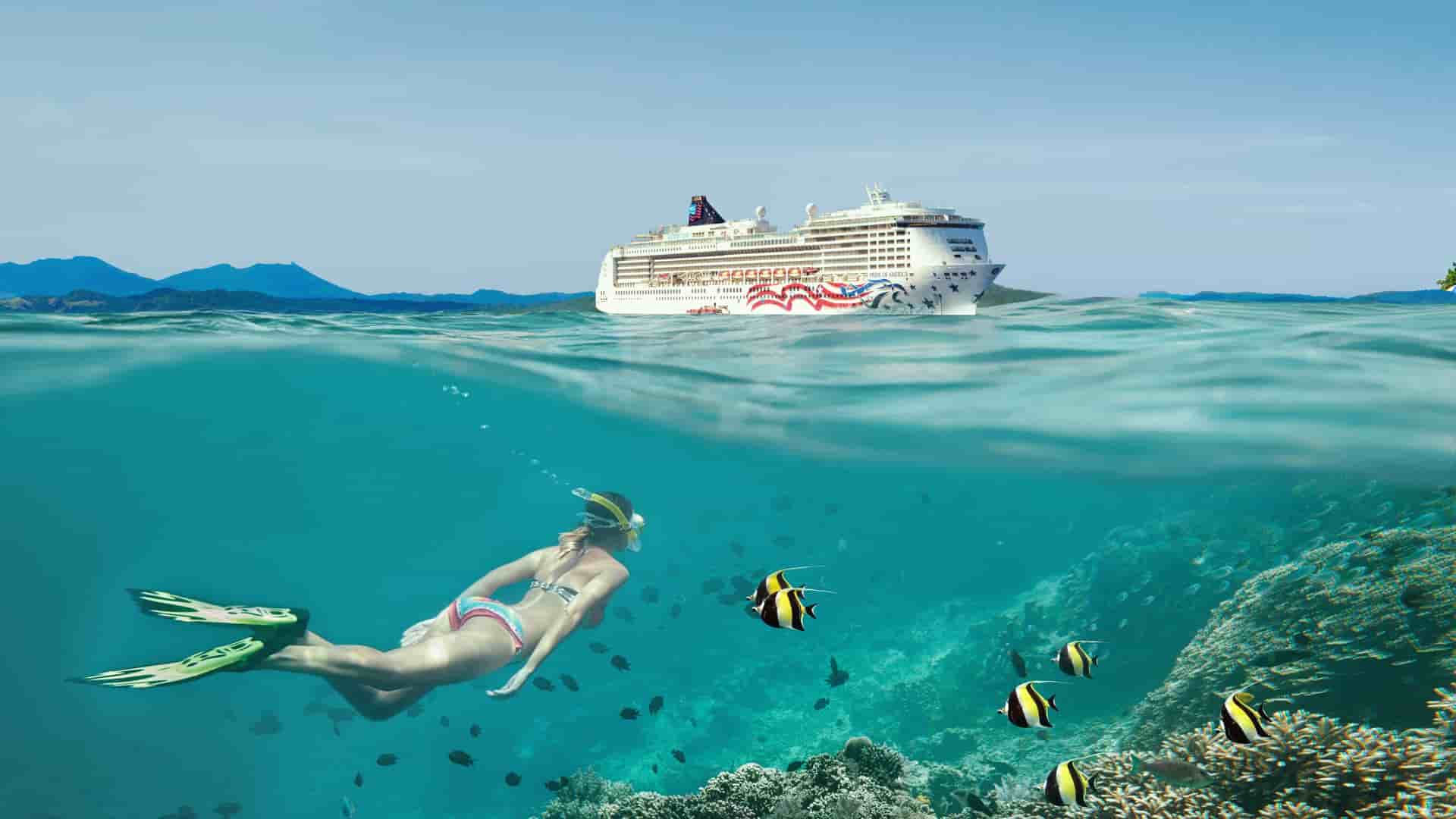 A snorkeler swims over a coral reef with tropical fish in clear Hawaiian water while the Pride of America cruise ship floats on the surface.