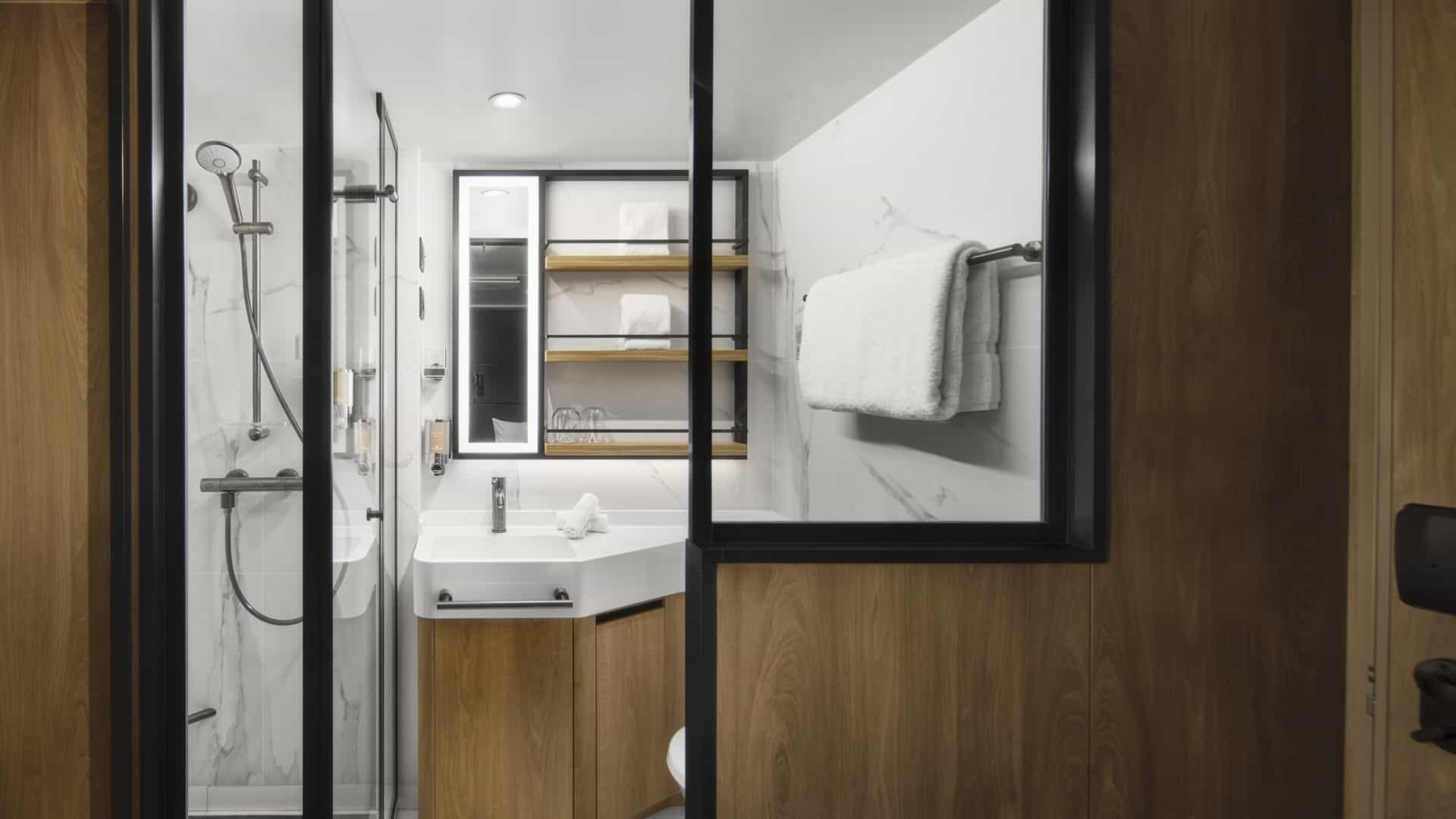 Bathroom in a Solo Studio on Norwegian Prima with a glass-enclosed shower, white sink and vanity, shelves and towels.