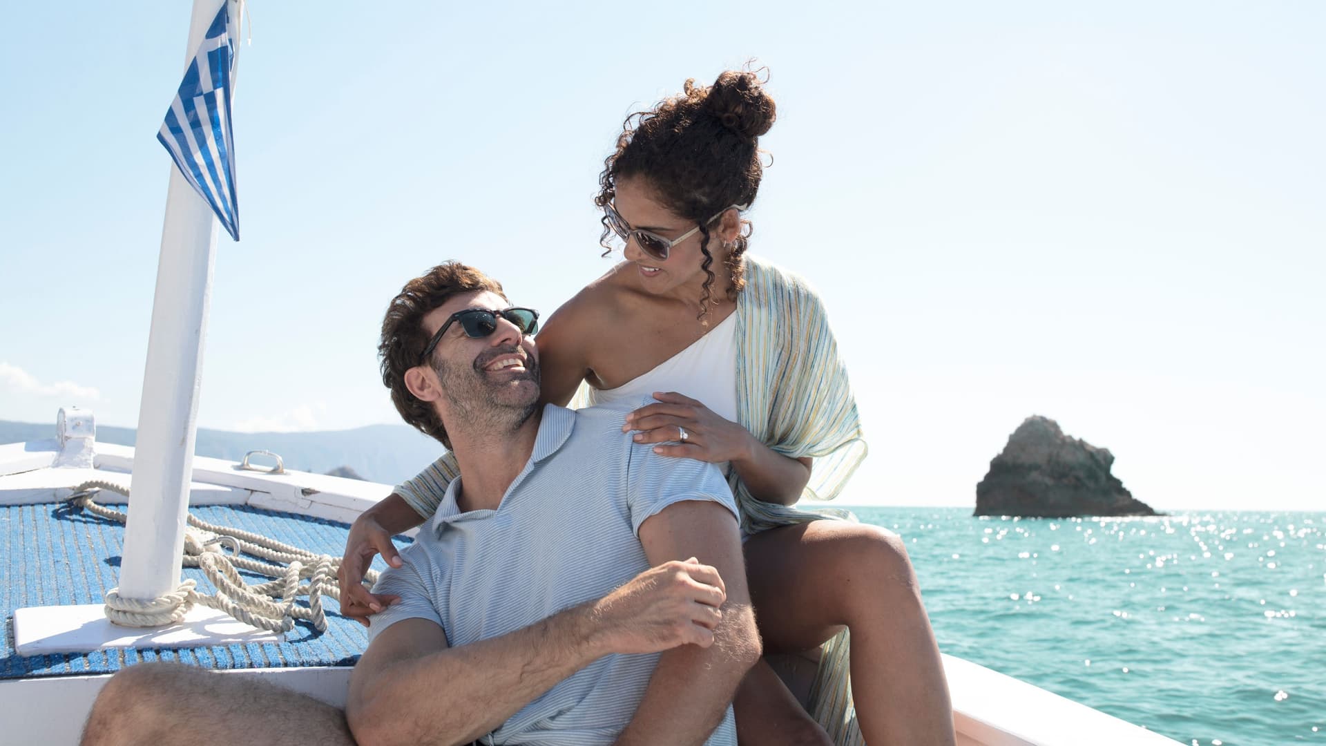 A happy couple smiles at each other during a sunny boat ride in Greece, with a small Greek flag visible on the bow and a rocky island rising from the turquoise sea in the background.