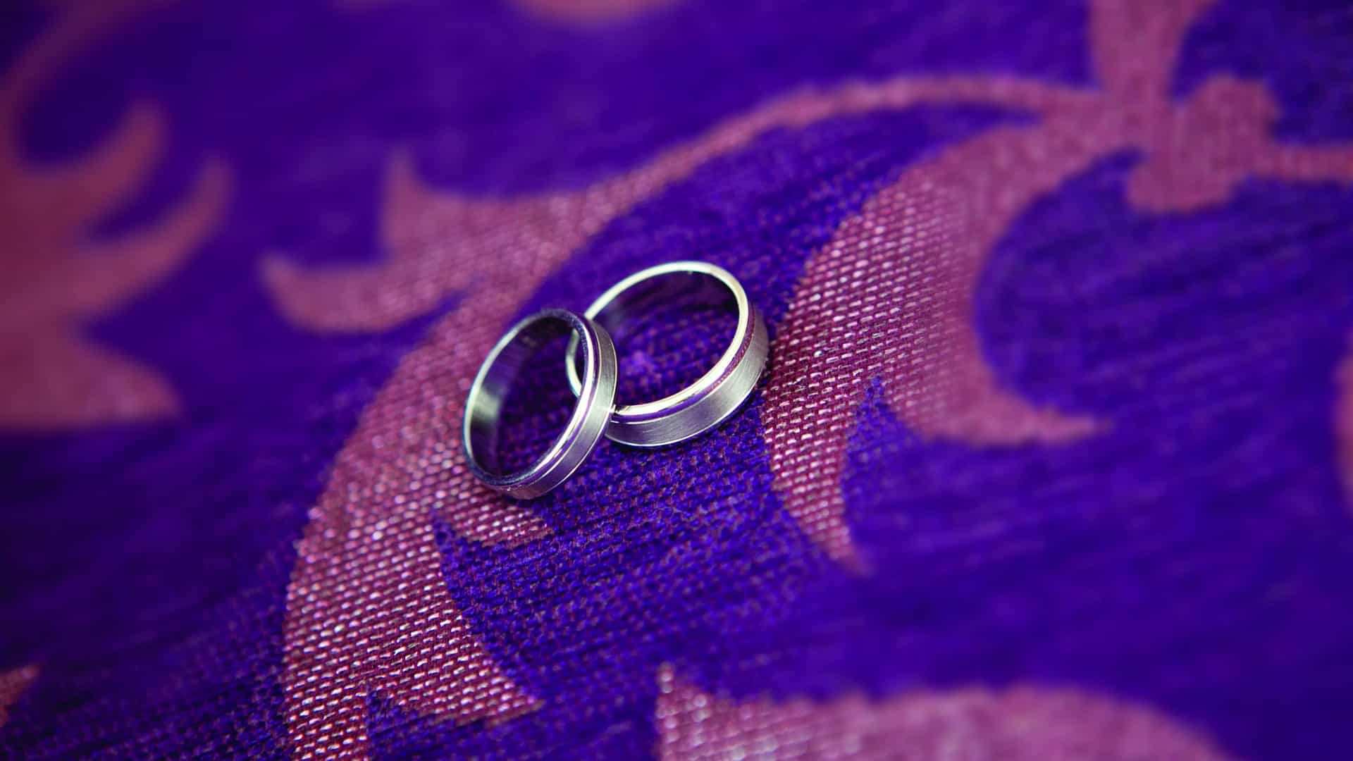 A close-up shot shows two silver wedding bands resting together on a textured, vibrantly patterned purple and pink fabric.