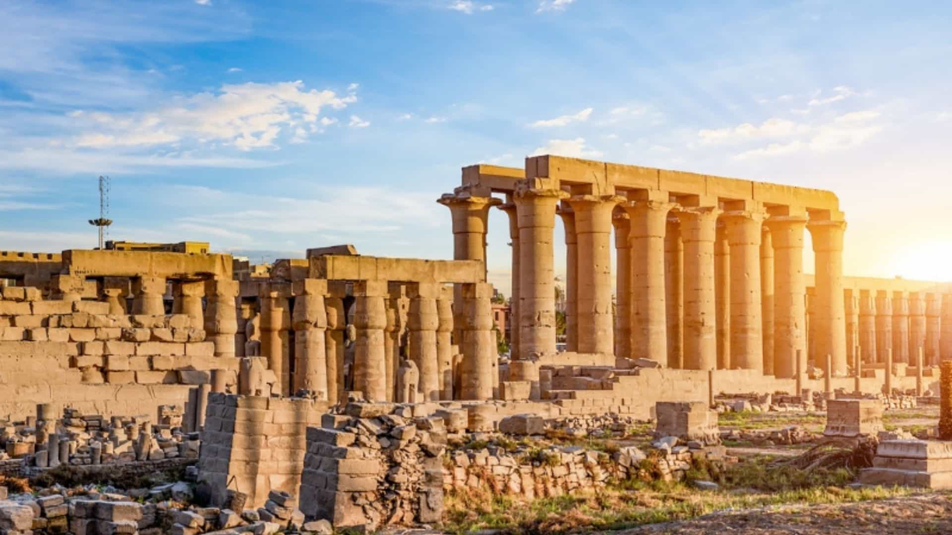 The ruins of the Karnak Temple in Luxor, Egypt, at sunset. The image shows the large, ancient stone columns and walls of the temple complex, which is located near the Nile River. The sun is setting on the right side of the image, casting a warm, golden glow on the stone structures and creating long shadows. The sky is bright blue with scattered white clouds, and the ground is covered in rubble and dry grass. The image beautifully captures the historical significance and dramatic light of this ancient Egyptian landmark.