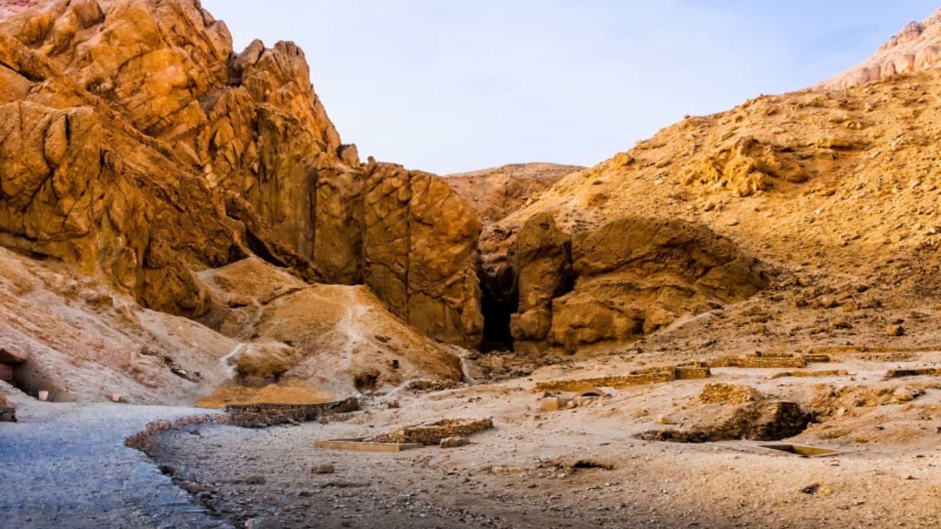 The entrance to the tomb of Tutankhamun, located in the rugged and arid landscape of the Valley of the Kings, Egypt. The image shows a narrow path winding through the valley's steep, rocky cliffs, with the dark entrance to the tomb visible at the center. The ground is a mix of sand, rock, and archaeological excavation sites. The sky is a bright blue, and the sun illuminates the dry, tan-colored rock formations. The scene captures the desolate but historically rich environment of this ancient Egyptian burial ground near the Nile River.