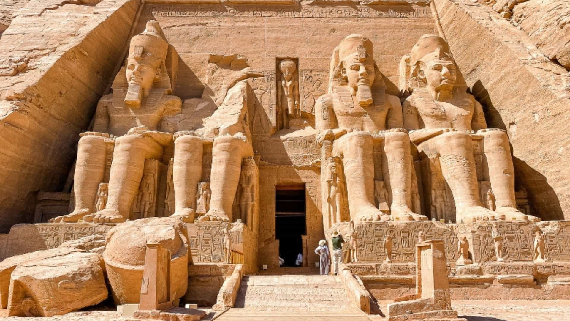 The Great Temple of Ramses II at Abu Simbel, Egypt. Four massive statues of the pharaoh, carved into a mountain, sit at the temple's entrance. The monumental statues are weathered and worn but still imposing. A wooden walkway leads to the entrance between two of the statues. Two people are visible at the base of the statues, providing a sense of scale. The temple is carved out of the reddish-brown rock, and hieroglyphs are visible on the facade. The image captures the grandeur and historical significance of this ancient Egyptian site near the Nile River.