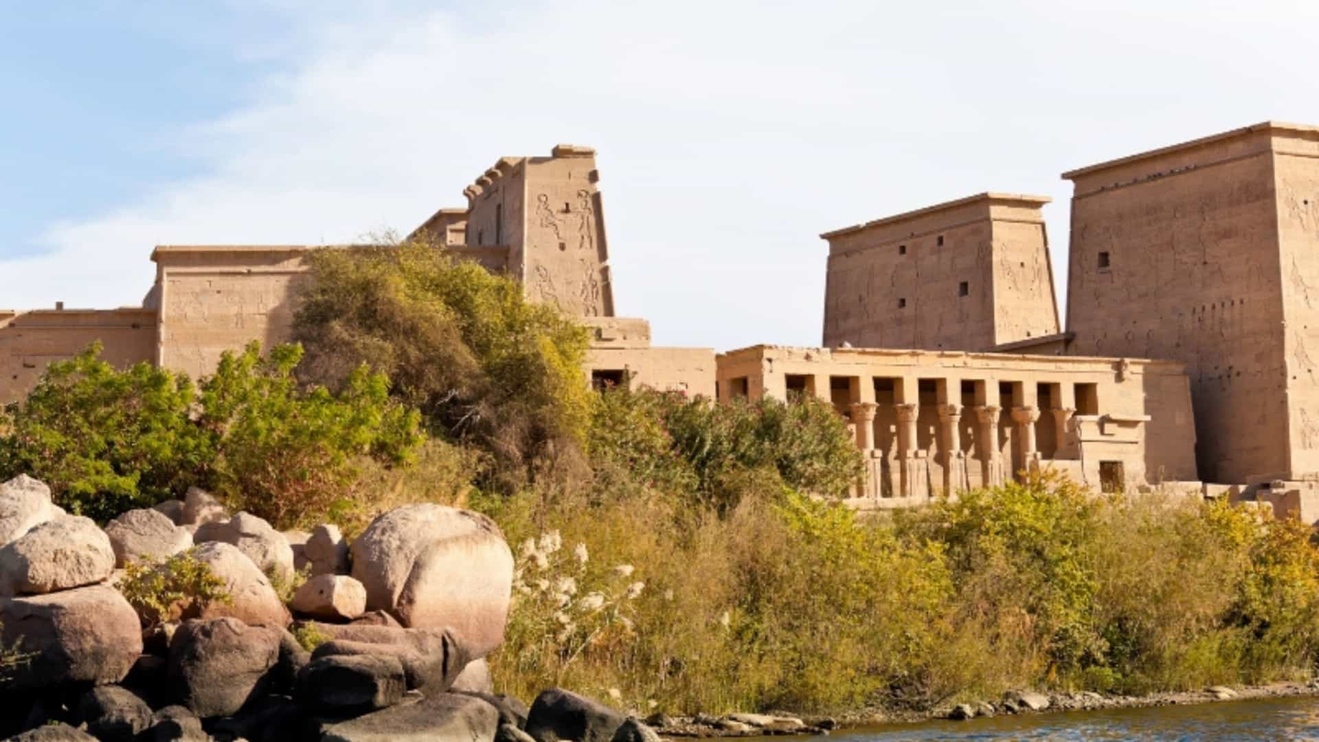 A view of the Philae Temple, dedicated to the goddess Isis, on an island in the Nile River in Egypt. The large, ancient stone structures, with their hieroglyphs and columns, rise above the riverbank, which is dotted with large rocks and green bushes. The water of the Nile is a deep blue in the foreground, and the sky is a bright blue with scattered clouds. The image captures the impressive architecture and serene setting of this ancient Egyptian temple.