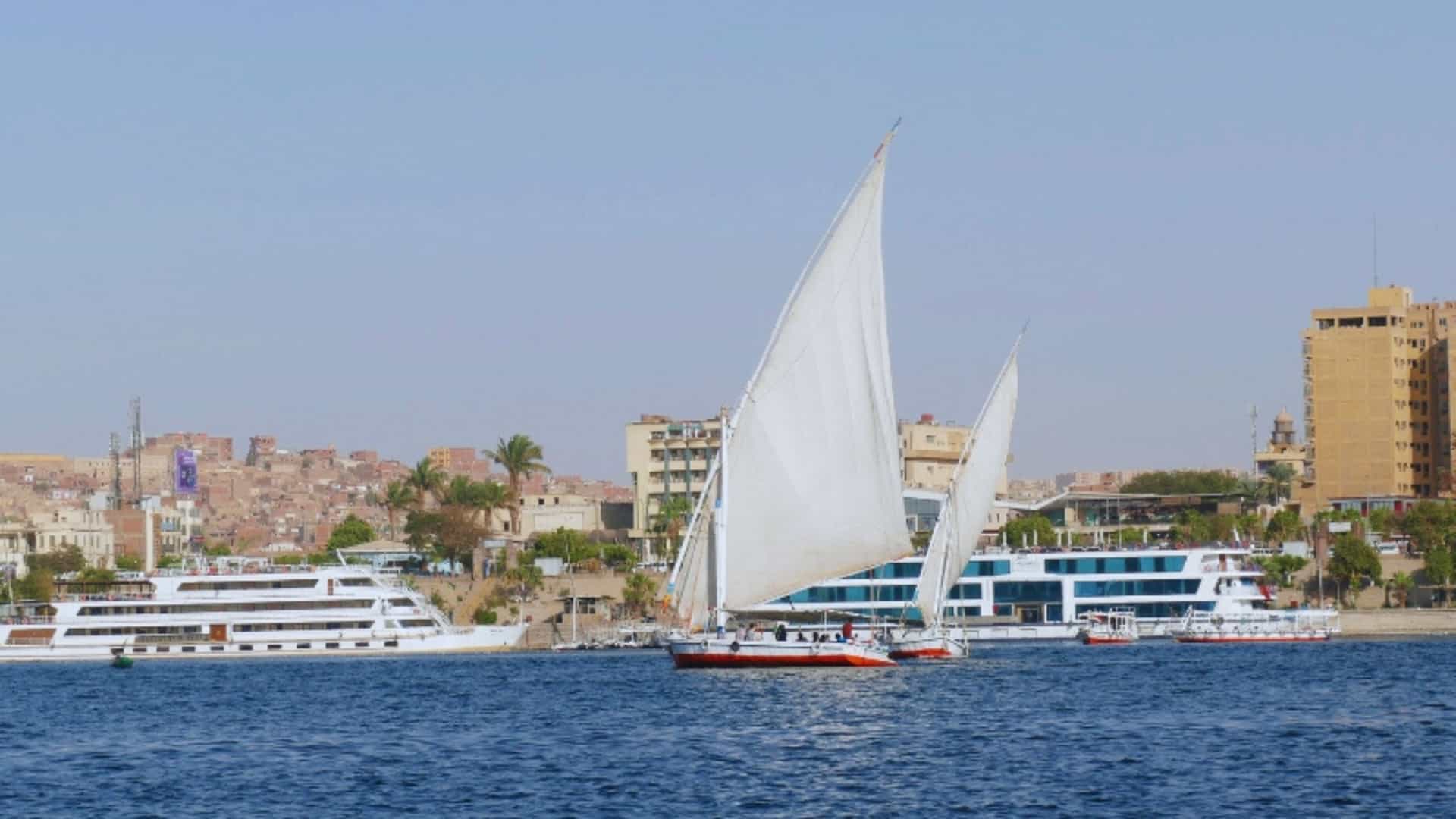 A traditional wooden felucca, with its distinctive triangular white sails, glides across the wide, blue Nile River in Egypt. The small boat is in the middle of the frame, passing by two large modern cruise ships docked at the riverbank. In the background, the cityscape of Aswan is visible, with multi-story buildings and a mix of brown and white structures. The sky is a clear, light blue, highlighting the contrast between ancient and modern forms of travel on this historic waterway.