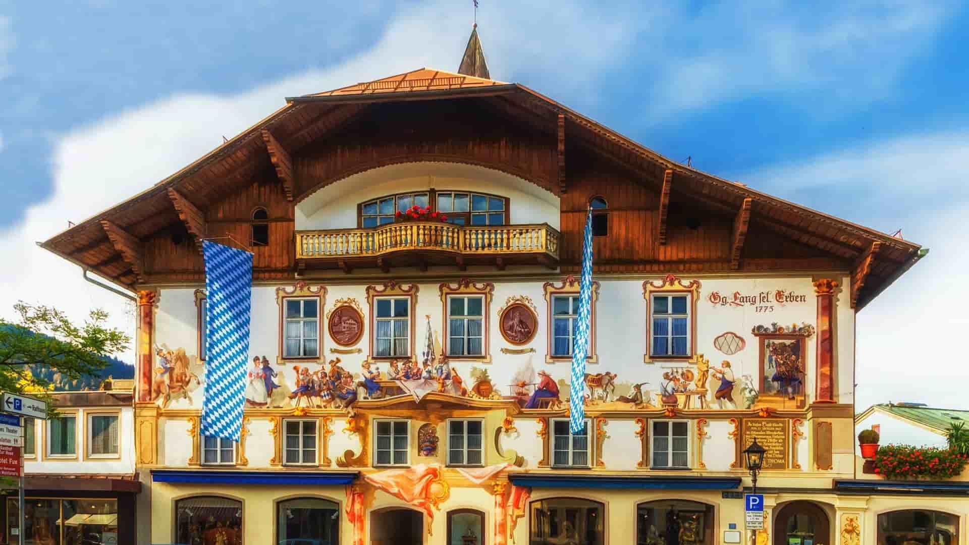 A traditional building in Oberammergau, Germany, showcases ornate exterior frescoes, a wooden balcony, and hanging blue-and-white Bavarian flags.