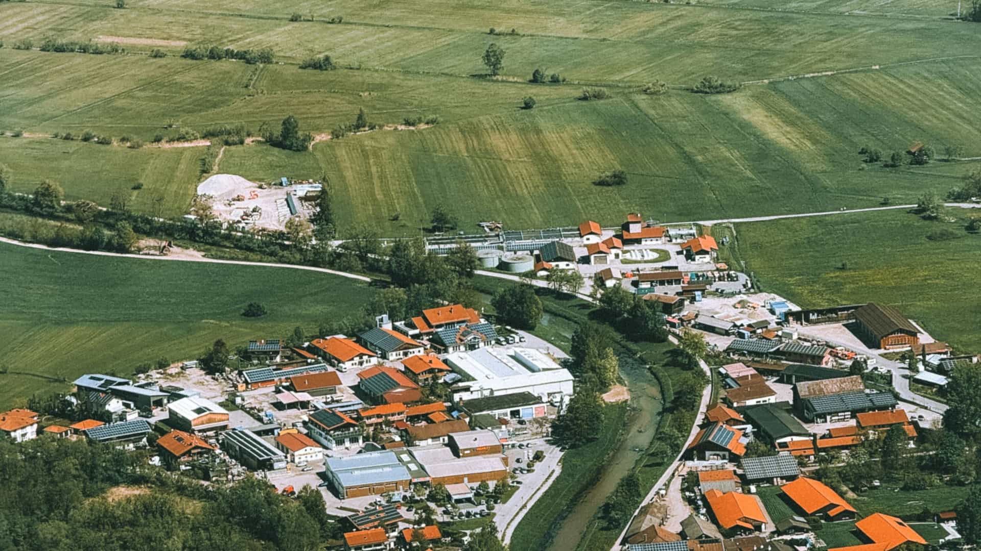 An aerial view shows a small rural village of Oberammergau, Germany, with red-roofed buildings nestled among green agricultural fields and a winding stream.