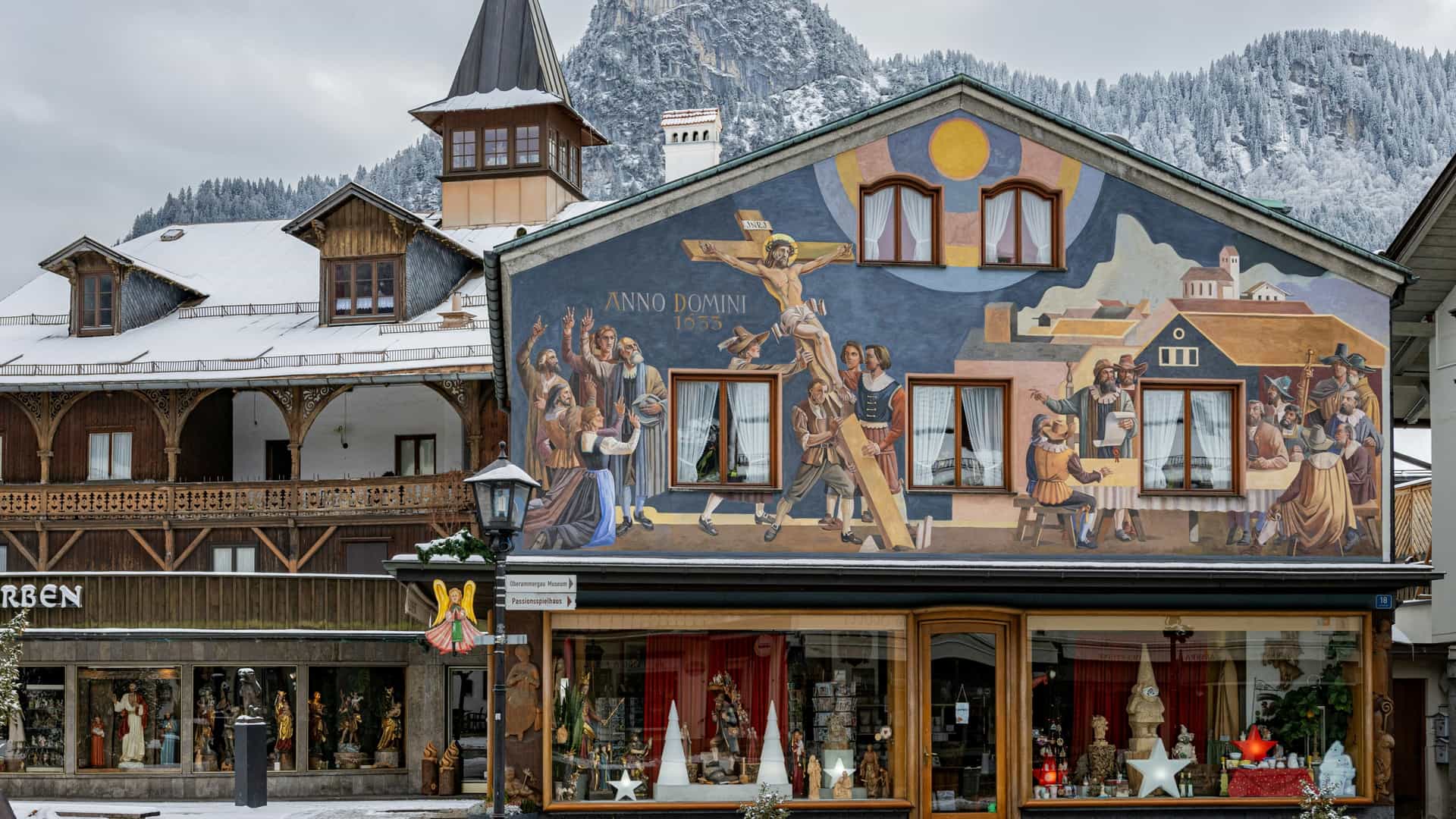 A traditional building in Oberammergau features a large exterior fresco depicting the 1633 historical scene of the Passion Play against a backdrop of snow-covered mountains and rooftops.