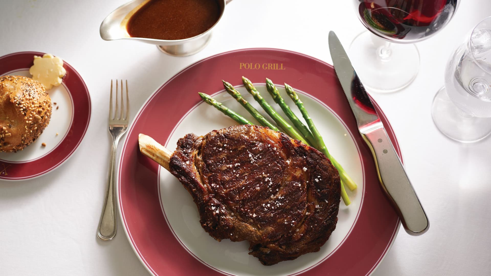 A bone-in ribeye steak with grilled asparagus and red wine sauce served on a Polo Grill branded plate aboard Oceania Cruises, accompanied by a sesame roll and red wine.