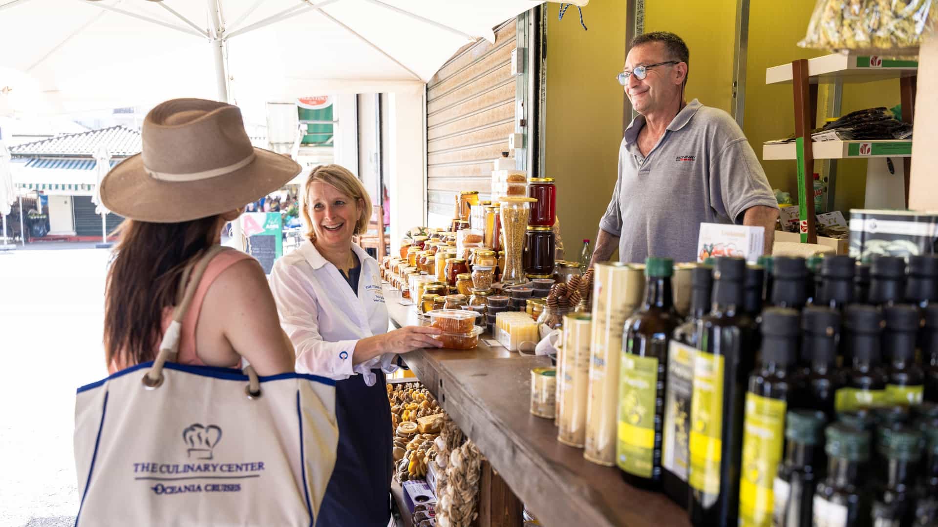 An Oceania Cruises Culinary Center chef instructor and guest browsing local honey, olive oil, and specialty products at an outdoor market stall during a Culinary Discovery Tour in Corfu, Greece.