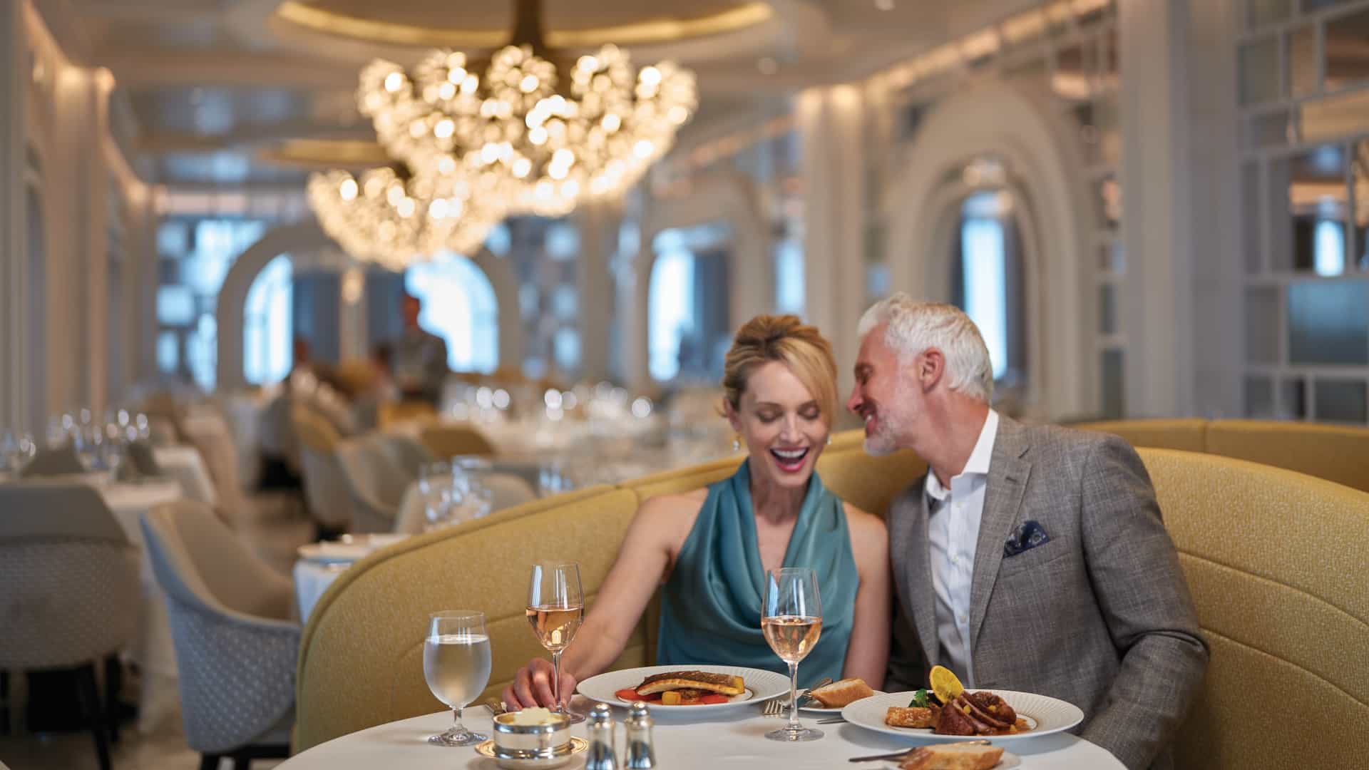 A couple enjoying dinner and wine in the elegantly appointed Grand Dining Room aboard an Oceania Cruises ship, surrounded by crystal chandeliers and white-linen tables.