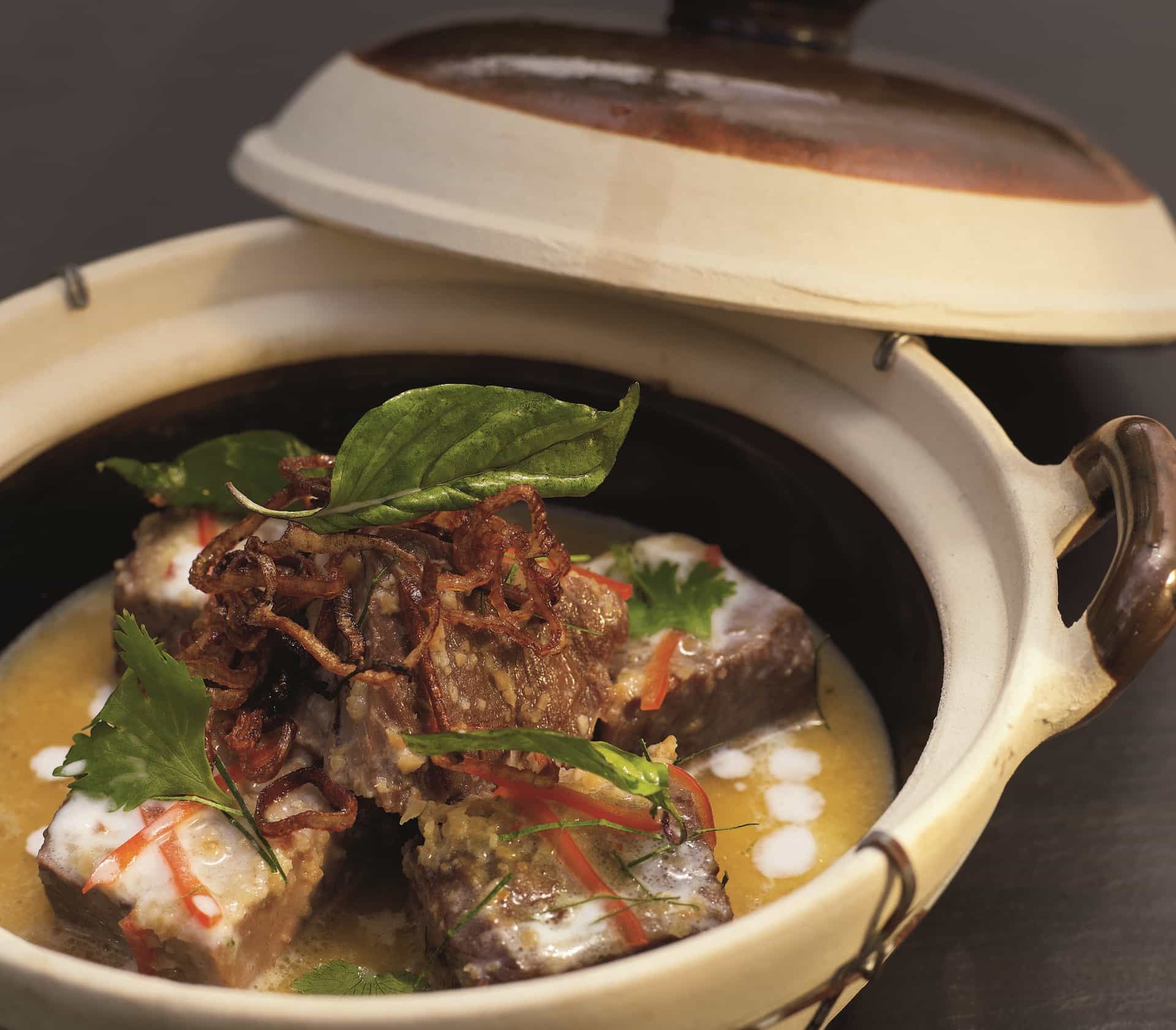 A braised lamb curry served in a ceramic pot with coconut broth, crispy shallots, fresh basil, chili, and herbs, as served in the Grand Dining Room on Oceania Cruises.