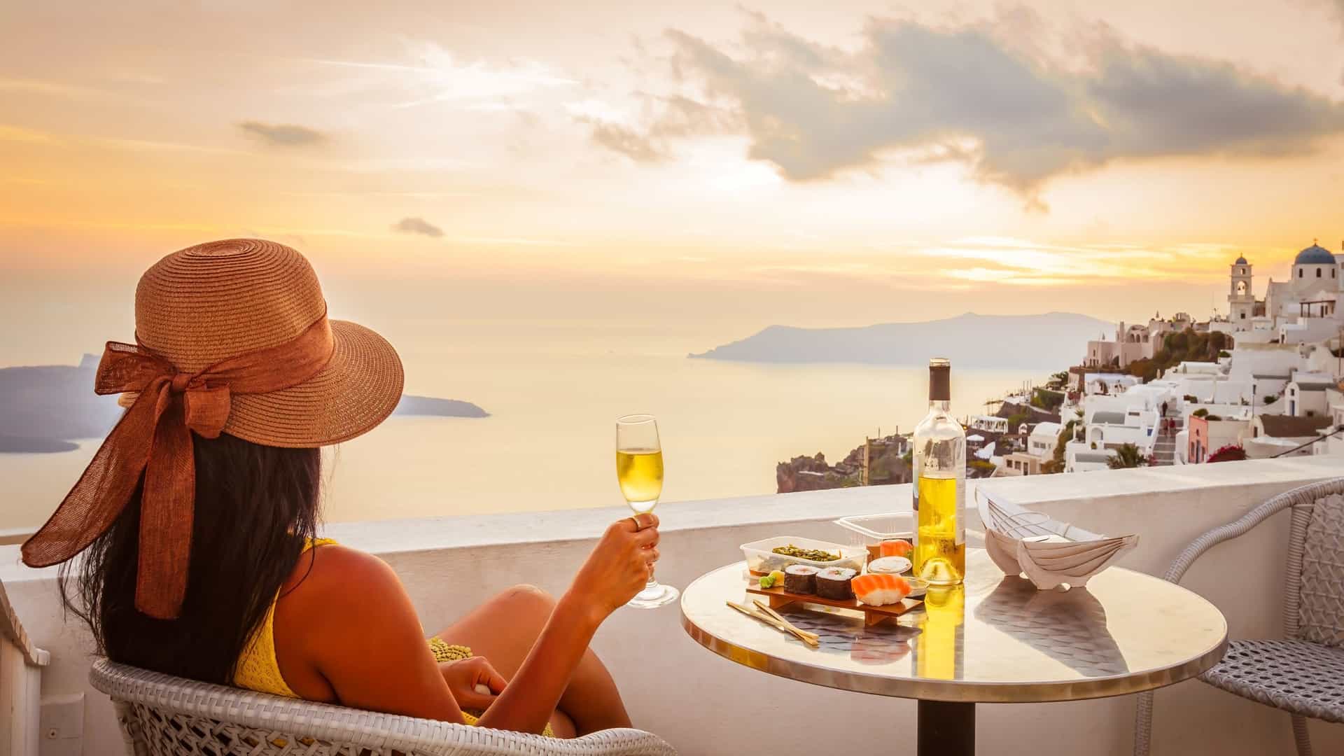 A traveler in a sunhat holds a glass of wine on a Santorini terrace overlooking whitewashed cliffside buildings and the Aegean Sea at sunset.