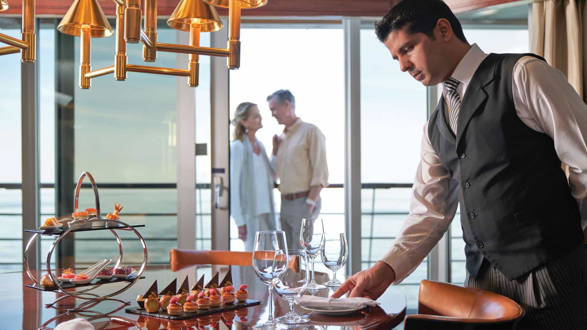 A butler in a dark vest and tie adjusts a setting at a dining table inside a luxury suite. In the background, an older couple stands by a large glass window looking out toward the ocean. A modern tiered serving stand filled with small canapés and a tray of pastries sit on the table in the foreground.