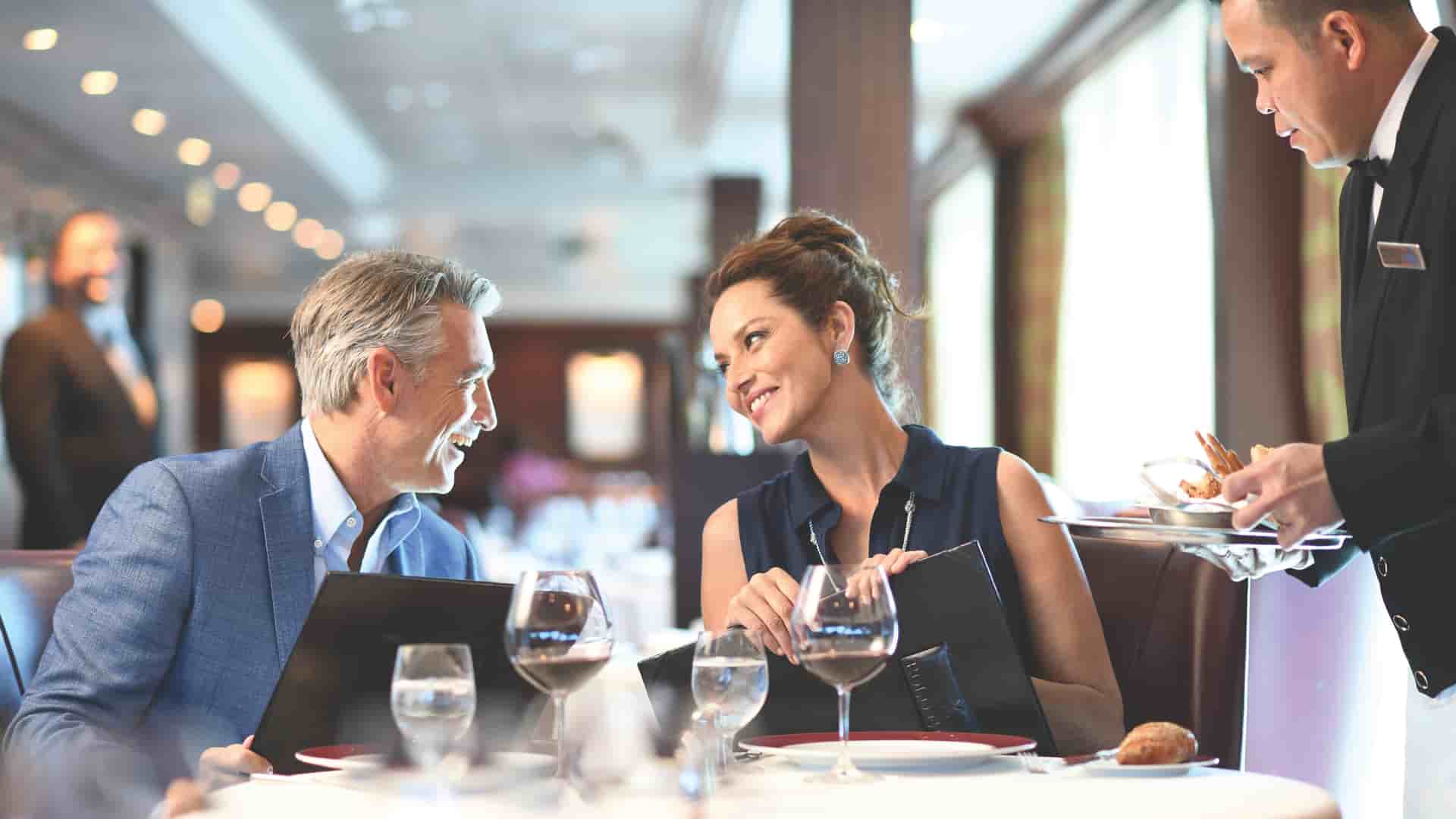 A couple sits at a table looking at each other while holding menus and getting ready to order with the waiter aboard Oceania Cruises in the Polo Gril Dining room.