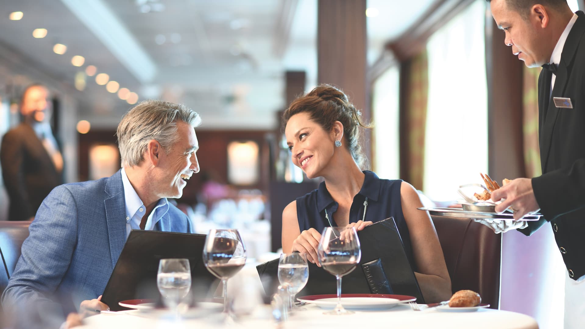 A couple sits at a table looking at each other while holding menus and getting ready to order with the waiter aboard Oceania Cruises in the Polo Gril Dining room.