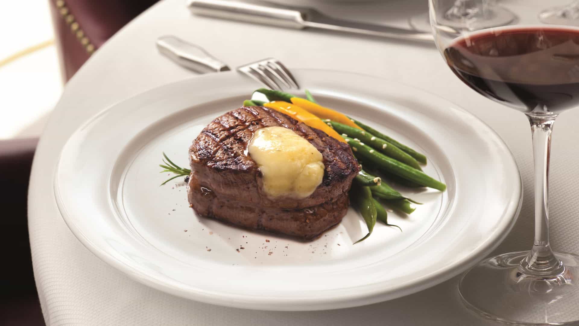 A grilled filet mignon topped with herb butter, served with green beans and yellow squash alongside a glass of red wine at Polo Grill on Oceania Cruises.
