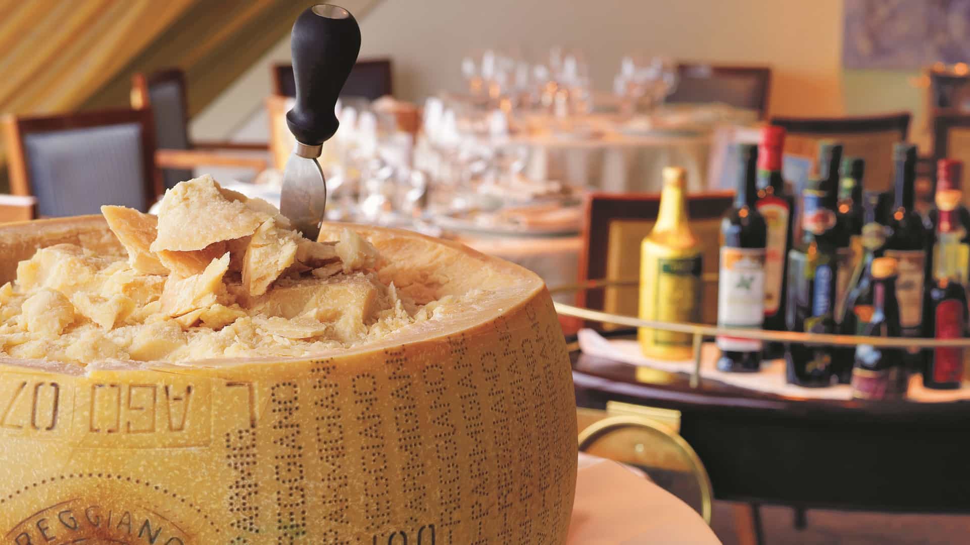 A close up of a wheel of parmesan cheese, with a fine dining tables and wine bottles blurred in the background.