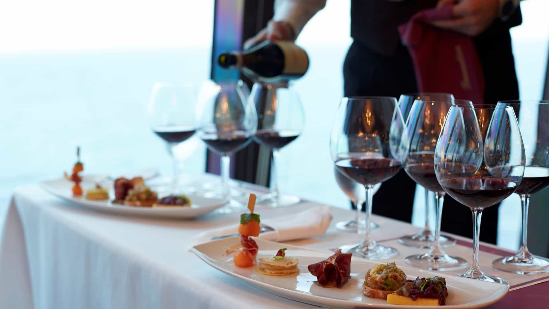 A sommelier pouring red wine alongside a curated selection of small plates and appetizers during an Oceania Cruises wine pairing event, with ocean views visible through the window.
