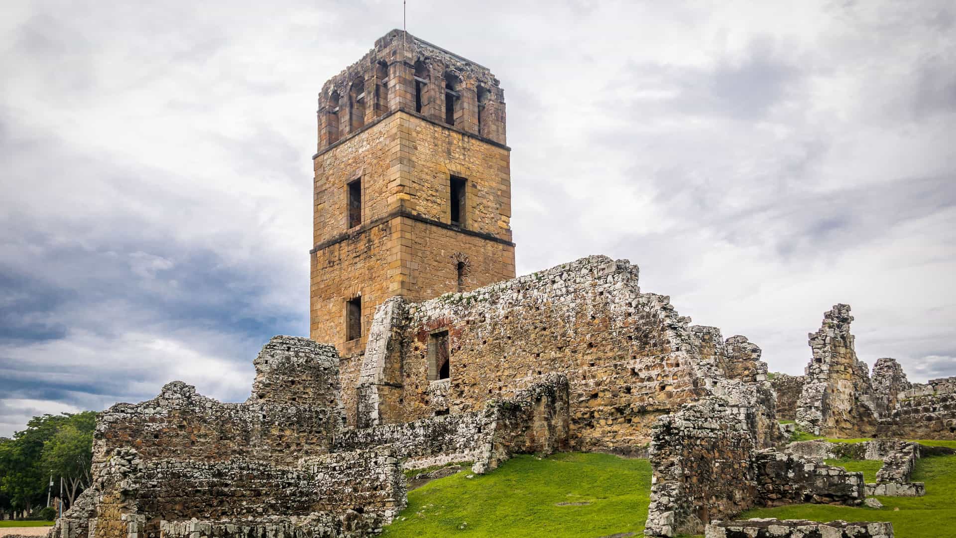 A dramatic view of the ruins of Panama Viejo in Panama City, a historically significant site near the Panama Canal. The tall, weathered stone tower of the old cathedral rises above the crumbling walls of the surrounding structures. The ruins sit on a grassy, slightly uneven field under a cloudy, dramatic sky. The image captures the historical importance and atmospheric feel of this ancient site.