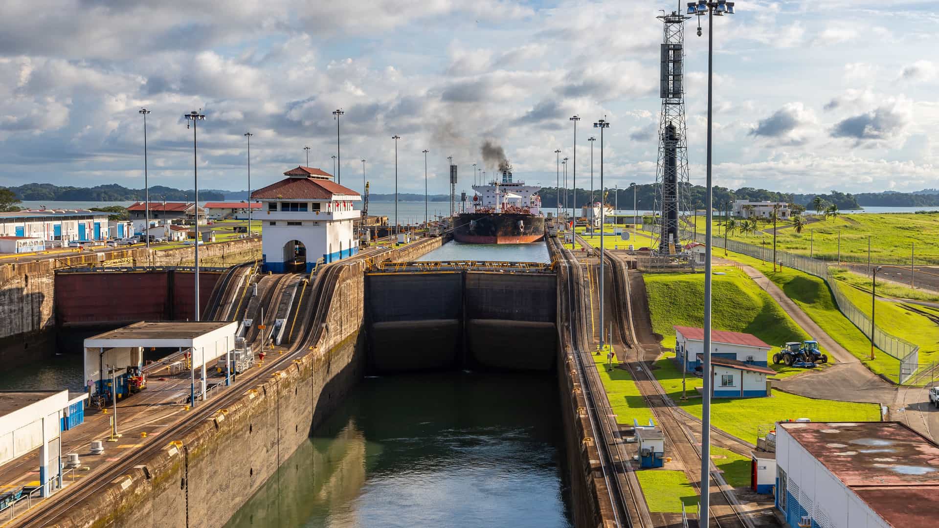 An elevated view of the Gatun Locks, a crucial part of the Panama Canal. A large cargo ship is positioned within one of the lock chambers, its deck loaded with containers. Water is visible on both sides, indicating the lock is in the process of raising or lowering the ship. Two other ships are visible in the distance on the lake. The locks are surrounded by green grassy areas and industrial buildings. The image captures the impressive scale and engineering marvel of this waterway.