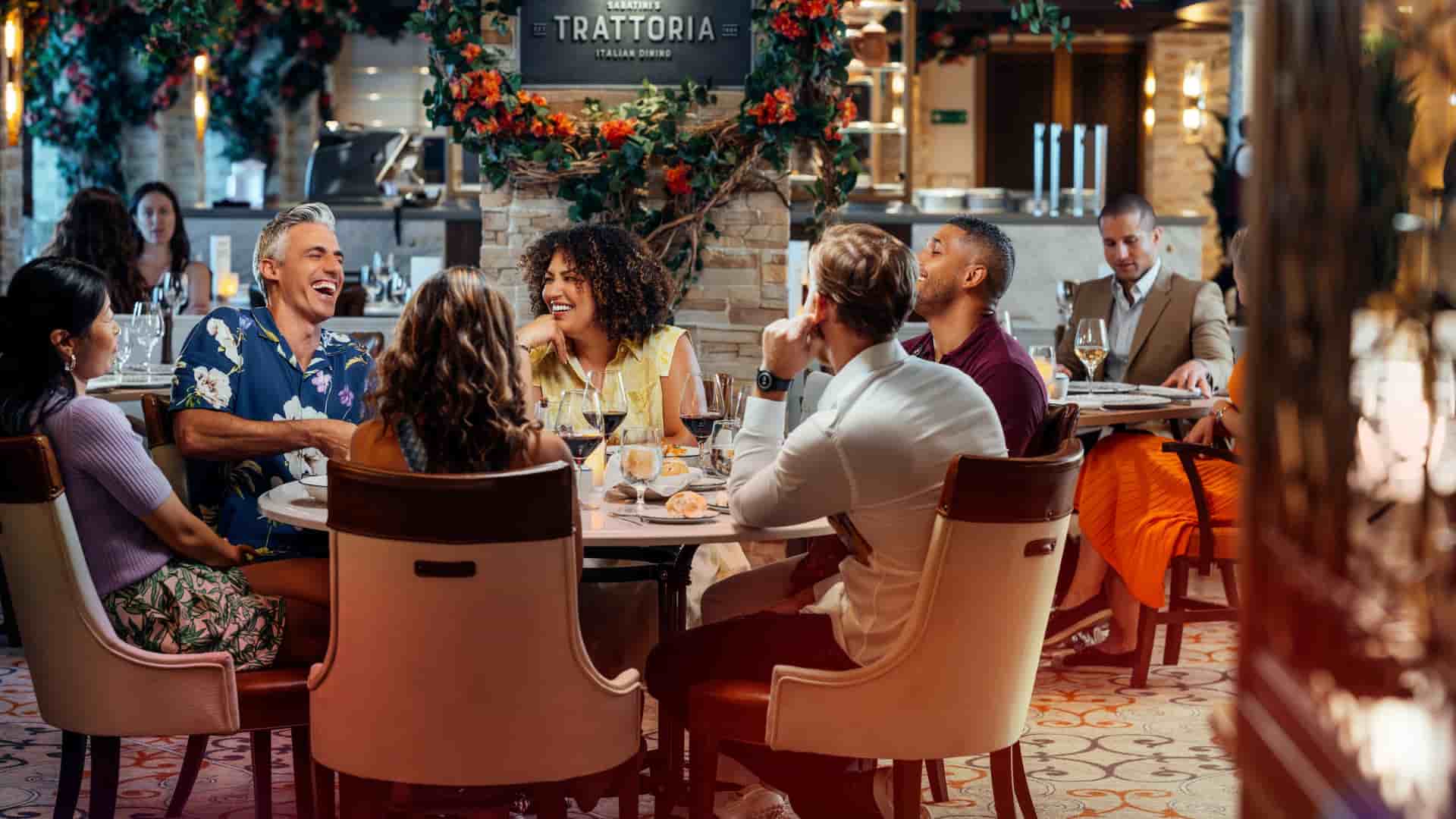 A diverse group of friends laughs and enjoys a meal together at a round table in the warm, rustic setting of Sabatini's Trattoria. The restaurant features elegant decor with stone pillars, floral garlands, and patterned carpeting that creates a vibrant, upscale atmosphere.