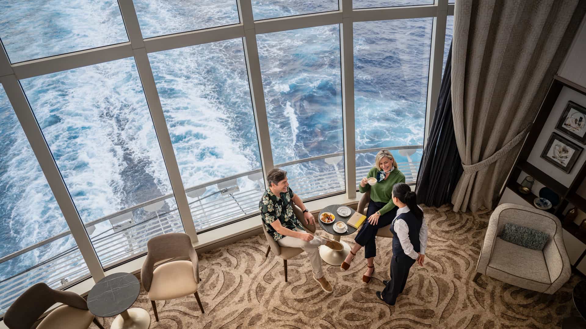 Aerial view of guests enjoying coffee and conversation with a crew member in the two-story Signature Lounge aboard Sun Princess, with floor-to-ceiling ocean views.