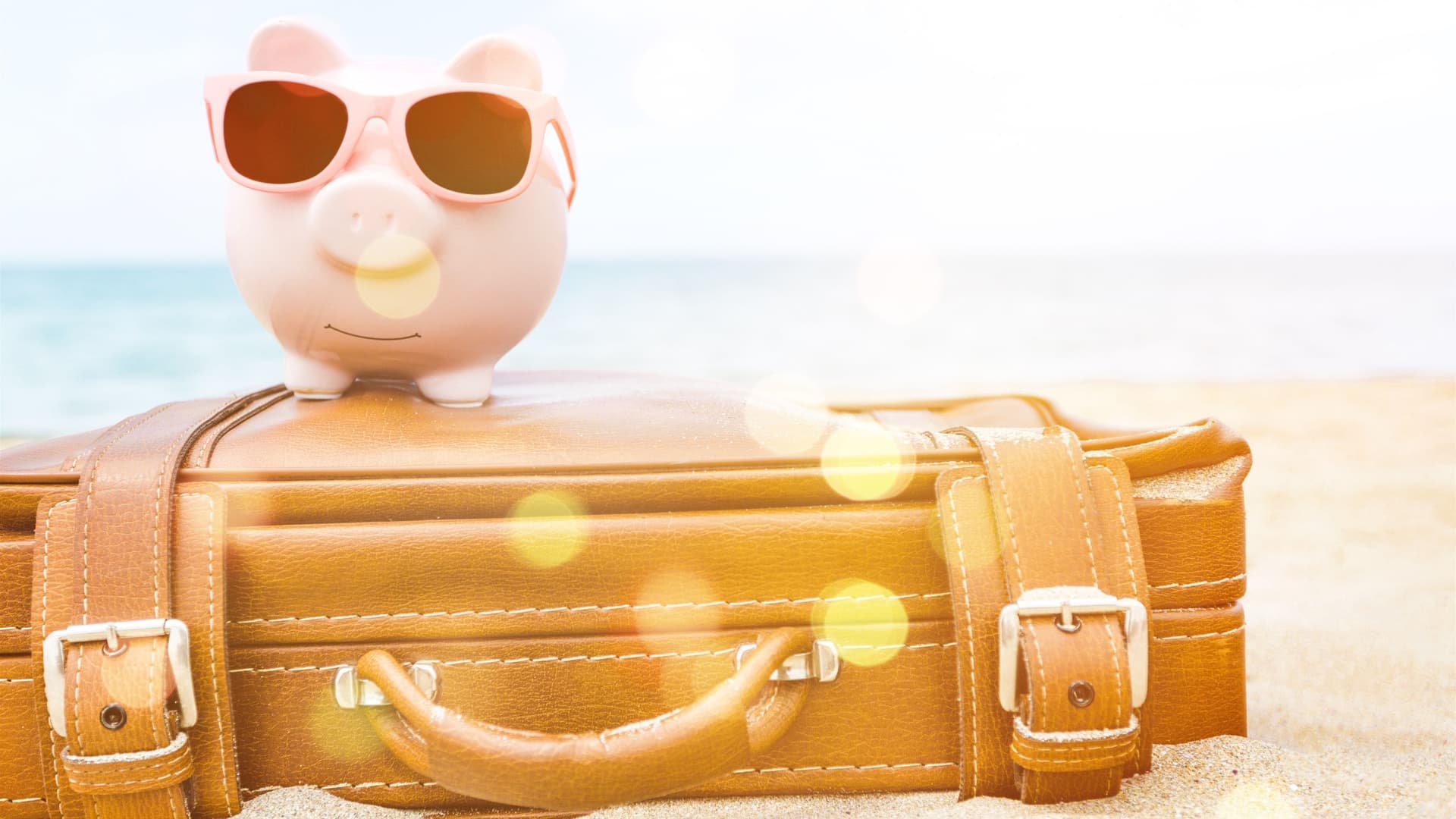 A piggy bank with sunglasses sits on top of a leather suit case on the shore of a beach.