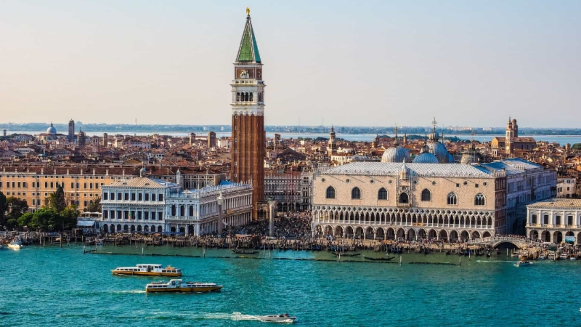 An iconic aerial view of Venice, Italy. St. Mark's Square is visible, with the famous Doge's Palace on the right, its facade of white marble and pink stone arches clearly visible. In the center, the tall, red-brick Campanile of St. Mark's Basilica rises toward a clear, bright sky. The wide, turquoise-blue canal is in the foreground, with several small boats and water taxis creating white wakes. The image captures the historical beauty of this city on the Po River Delta.