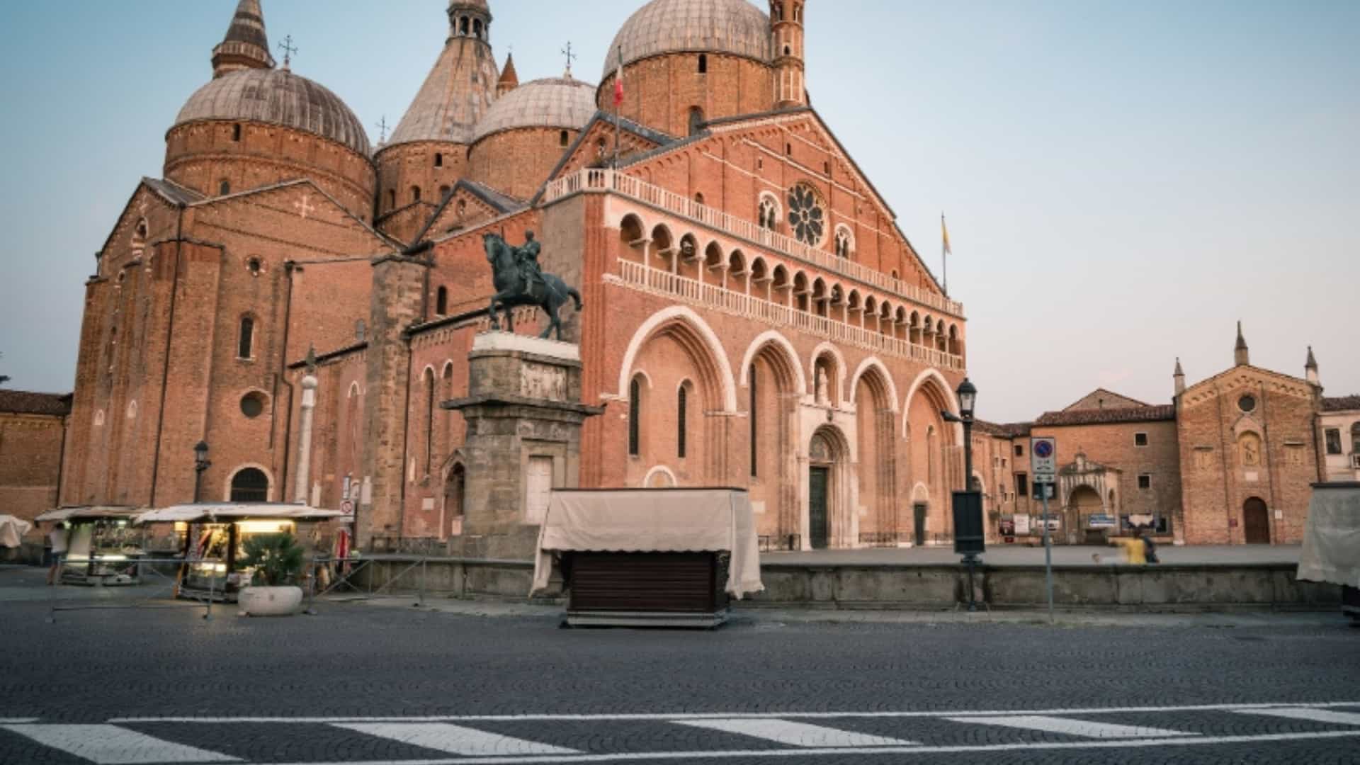 The Basilica of Saint Anthony of Padua in Italy, a large Roman Catholic church with distinctive domes and brick facade. A statue of a man on a horse is in front of the main entrance, and the ground is covered in cobblestones. The image captures the historical architecture of the Po River region.