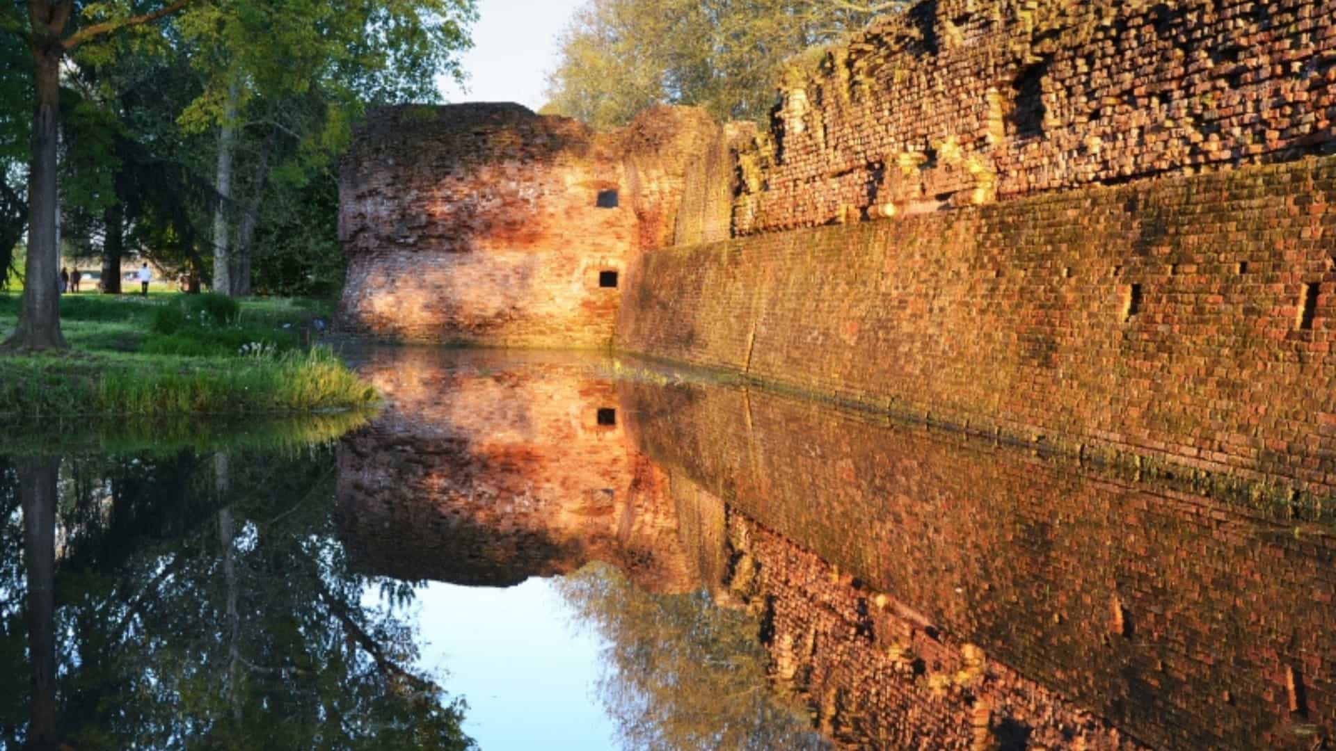 The ancient red-brick walls and a moat of a medieval castle in Ferrara, Italy. The walls are partially in shadow while the sun shines on the upper section, reflecting clearly on the calm water below. The image highlights the historic architecture found near the Po River.