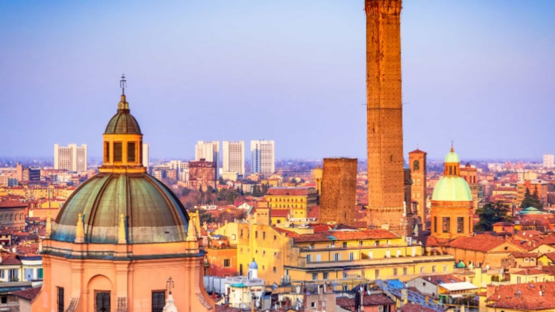 A panoramic view of the Bologna skyline in Italy, a city in the Po River valley. The image features a cluster of historic buildings with red-tiled roofs and a prominent medieval tower rising above them. The sky is a gradient of blue and orange, and the cityscape showcases the unique architecture of the Po River region.
