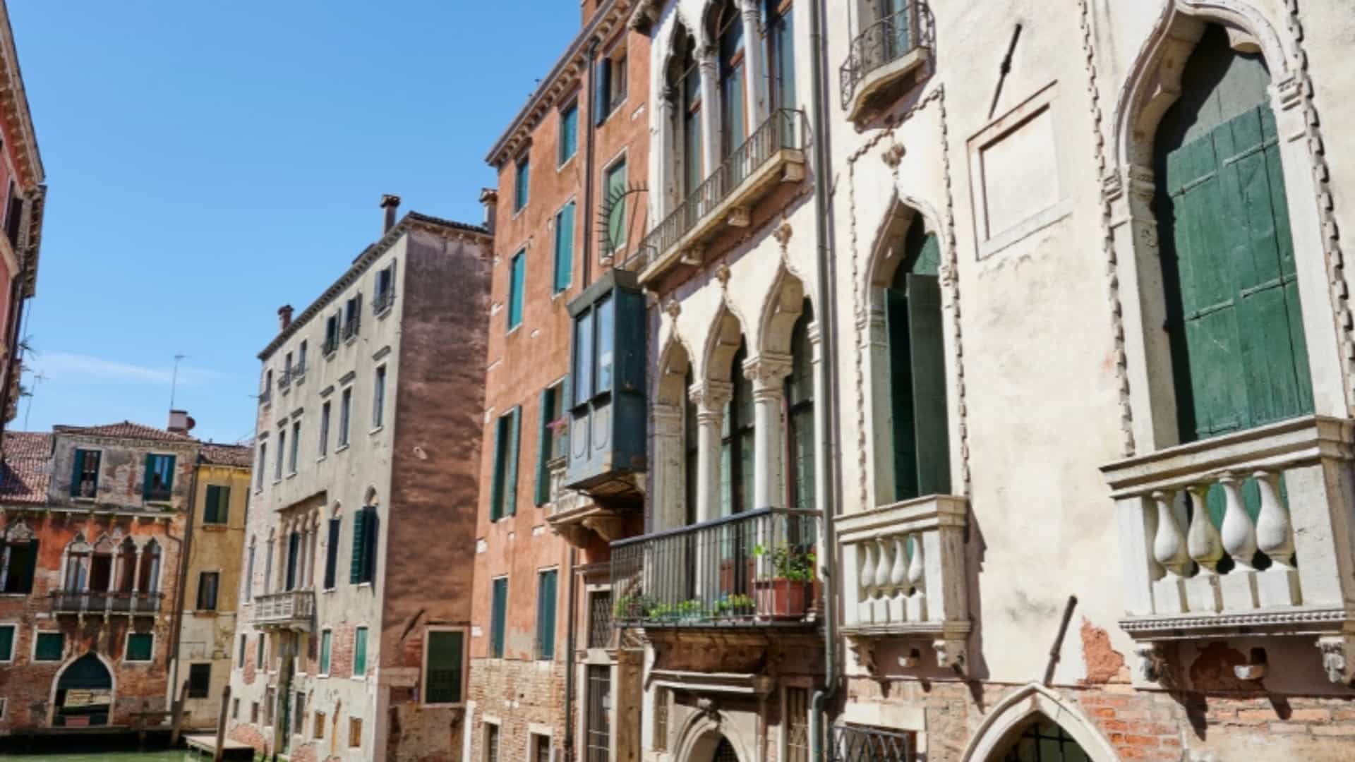 A view of a canal in Venice, Italy, surrounded by historic buildings with aged facades, intricate windows, and small balconies. The water in the canal reflects the buildings. The image captures the unique city architecture and waterways found in the Po River Delta region.