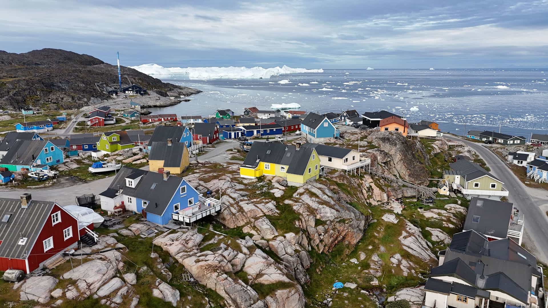 A wide-angle aerial shot of the remote Aappilattoq village in Greenland, with vibrant, colorful houses built on rocky terrain overlooking the Prince Christian Sound filled with ice floes and large icebergs.