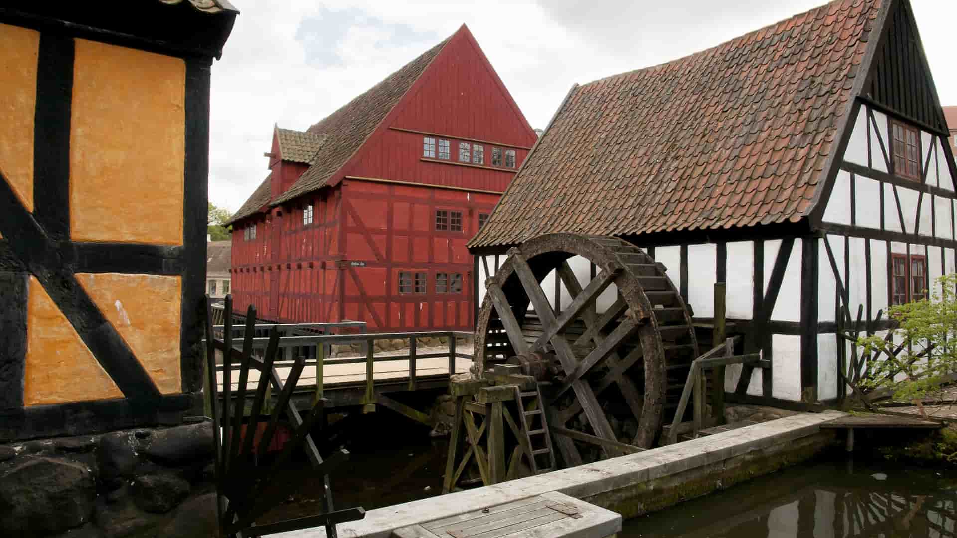 A vintage half-timbered building with a large wooden water wheel in a stream, part of the open-air Den Gamle By museum in Aarhus, Denmark.