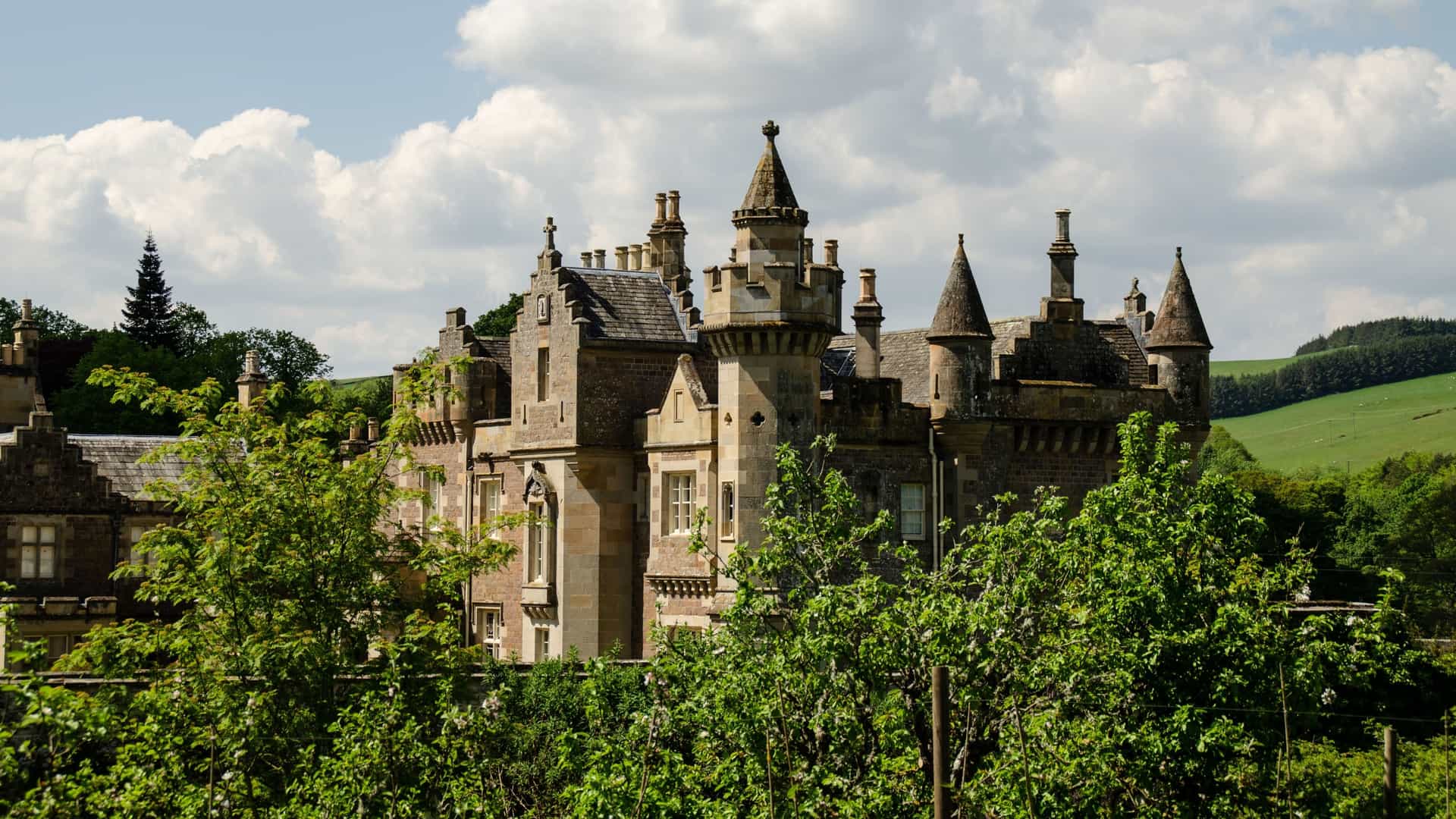 A historic, gothic-style country house with turrets and chimneys, the Abbotsford House, home of Sir Walter Scott in the Scottish Borders, surrounded by lush green trees and rolling hills.