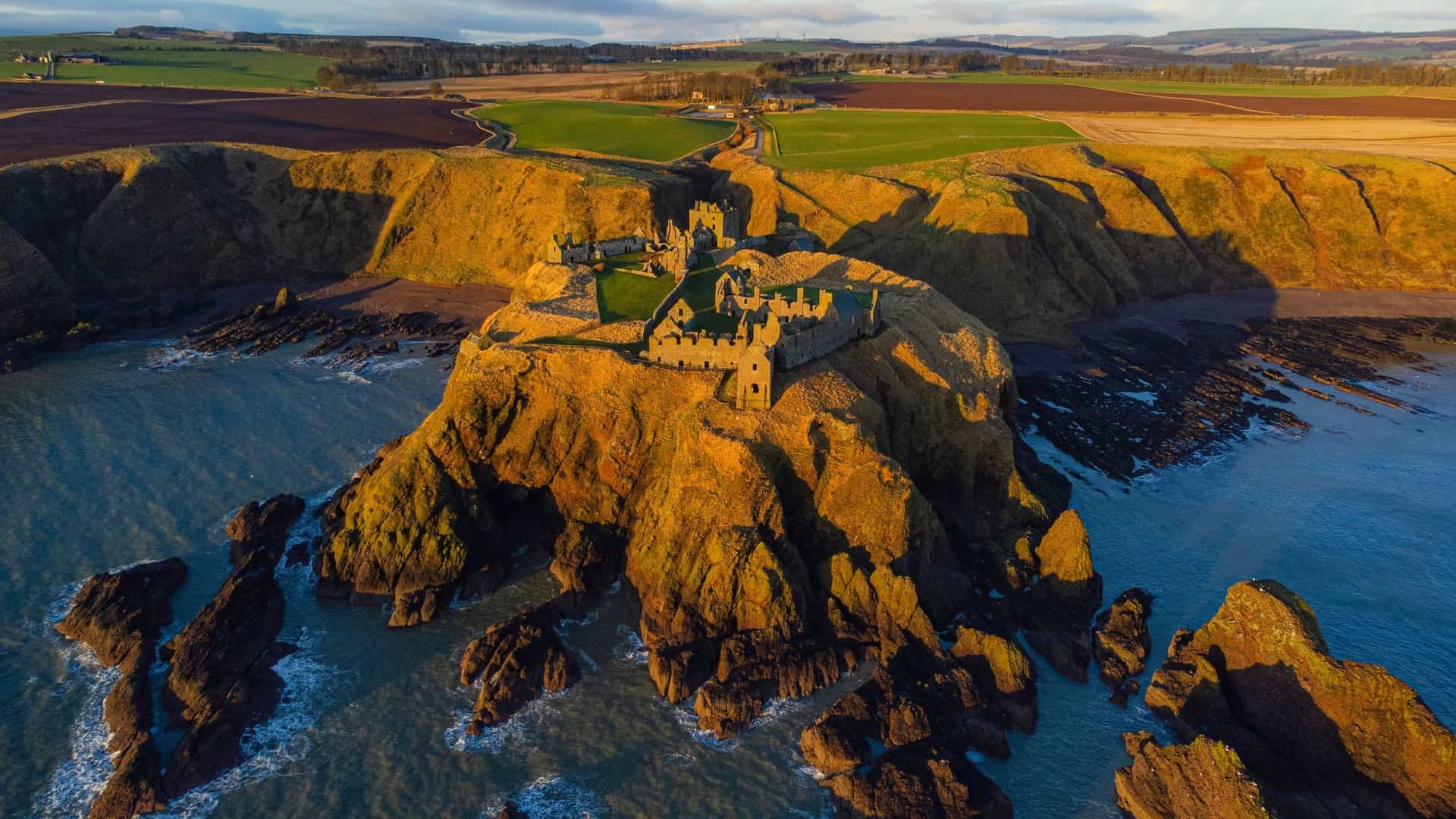 An aerial view of the dramatic ruins of Dunnottar Castle perched on a cliffside promontory overlooking the sea, a historical landmark near Aberdeen, Scotland.