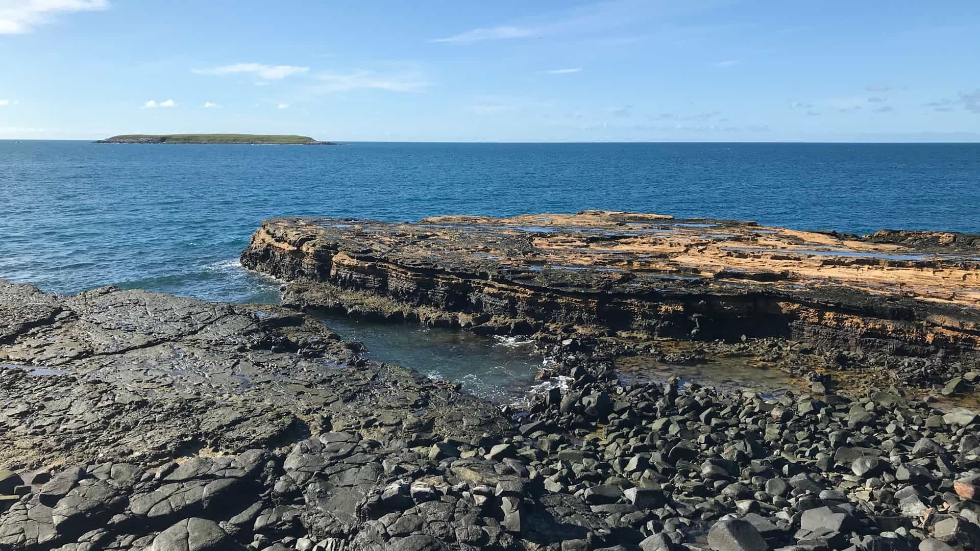 A scenic view of the Abrolhos Archipelago National Park, with a rocky coastline in the foreground and a small island in the vast blue ocean under a clear sky.