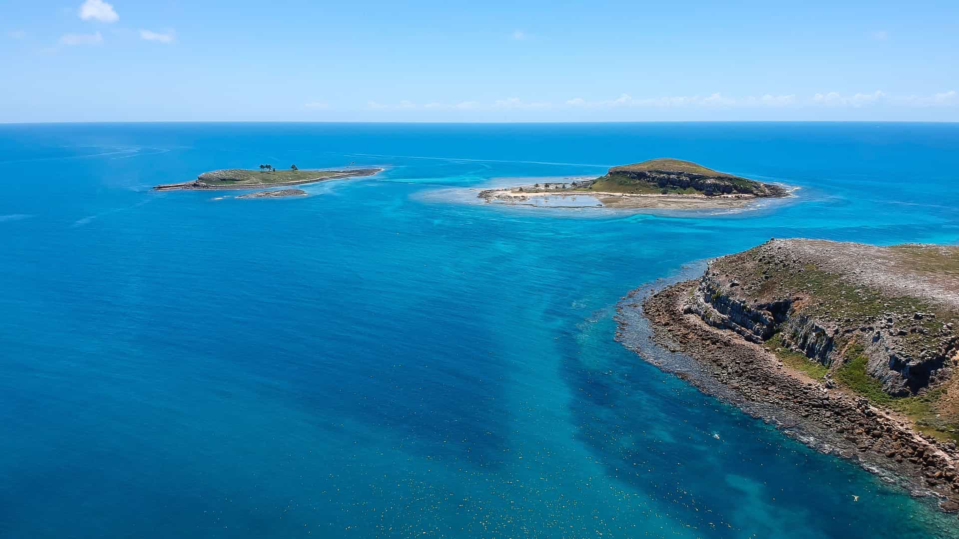 An aerial view of the Abrolhos Islands, with multiple small islands surrounded by crystal-clear turquoise water and reefs under a bright blue sky.