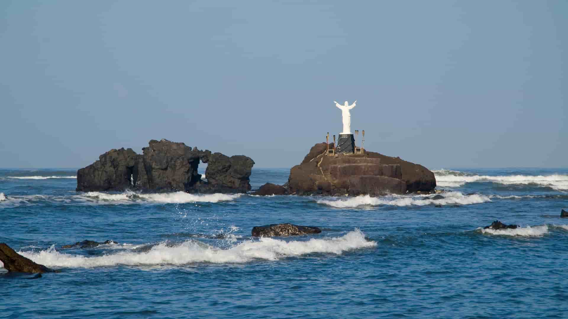 A statue of Jesus stands on a rocky islet in the Pacific Ocean, with waves crashing around it near the port of Acajutla, El Salvador.