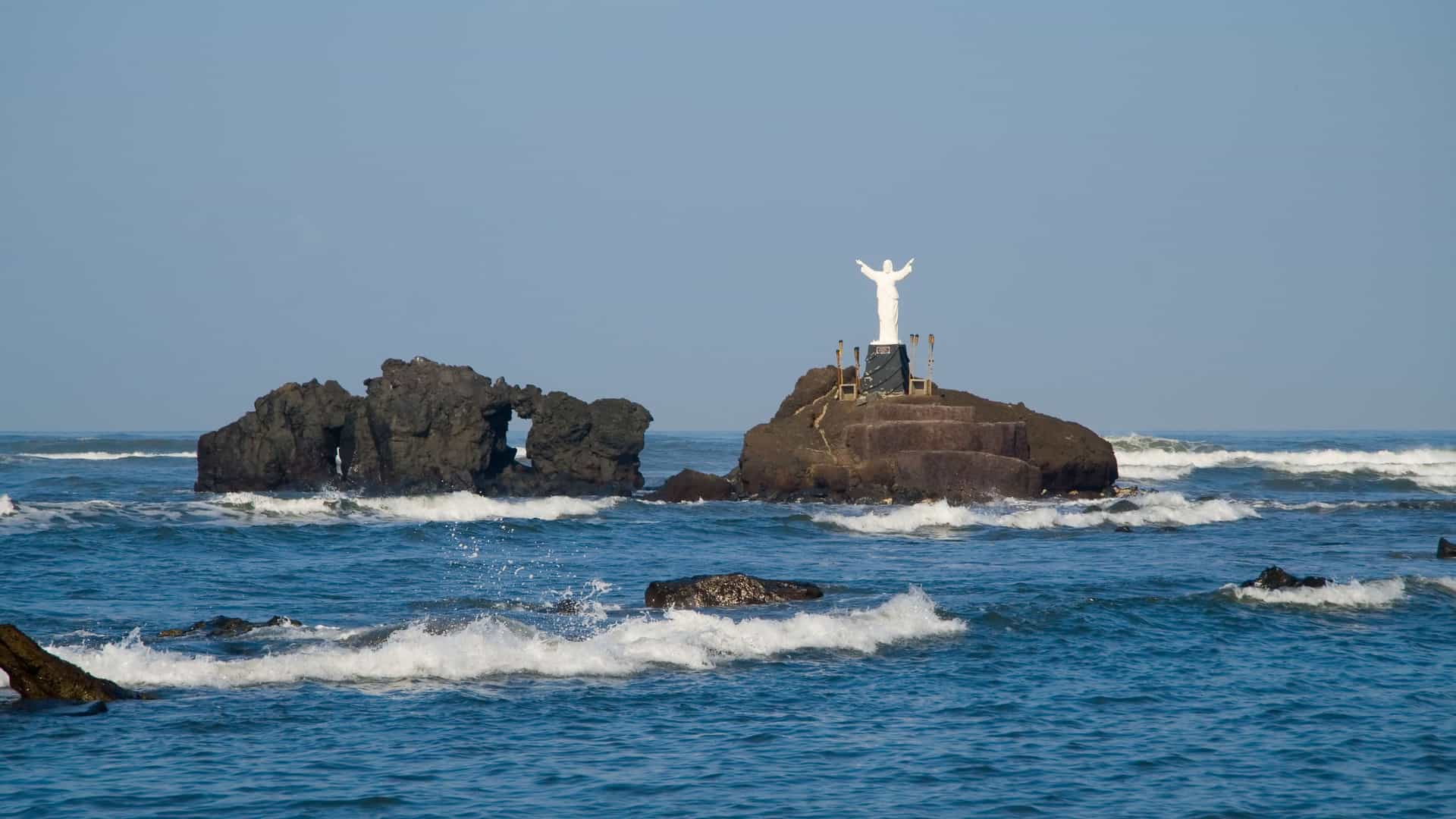 A statue of Jesus stands on a rocky islet in the Pacific Ocean, with waves crashing around it near the port of Acajutla, El Salvador.