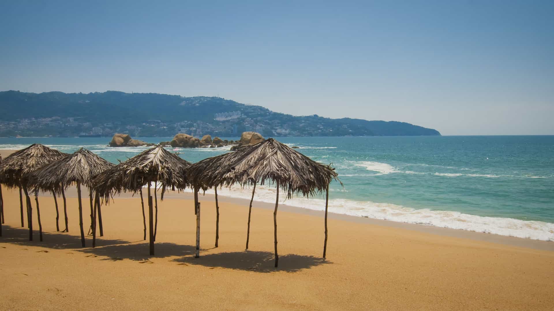 A beautiful beach in Acapulco, Mexico, with thatched-roof umbrellas on the golden sand, facing the turquoise ocean under a clear blue sky.