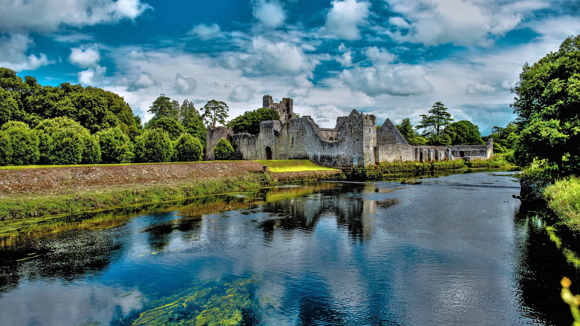 A scenic view of the stone ruins of the Adare Friary on the bank of the River Maigue in Adare, Ireland. The large, weathered ruins are surrounded by lush green trees, and the calm water of the river in the foreground reflects the dramatic blue and white cloudy sky.
