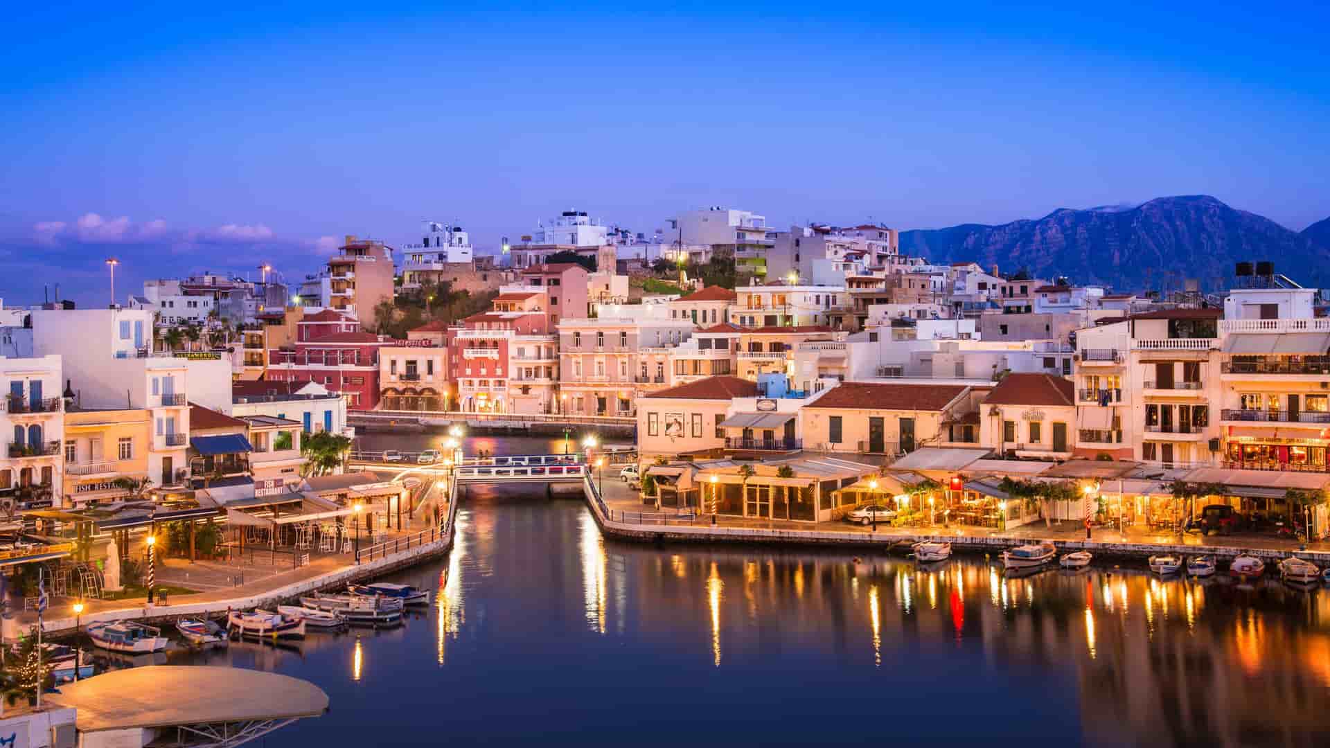 A stunning dusk shot of Agios Nikolaos, Crete, with colorful buildings and restaurants lining the tranquil Voulismeni Lake, their lights reflecting on the water.