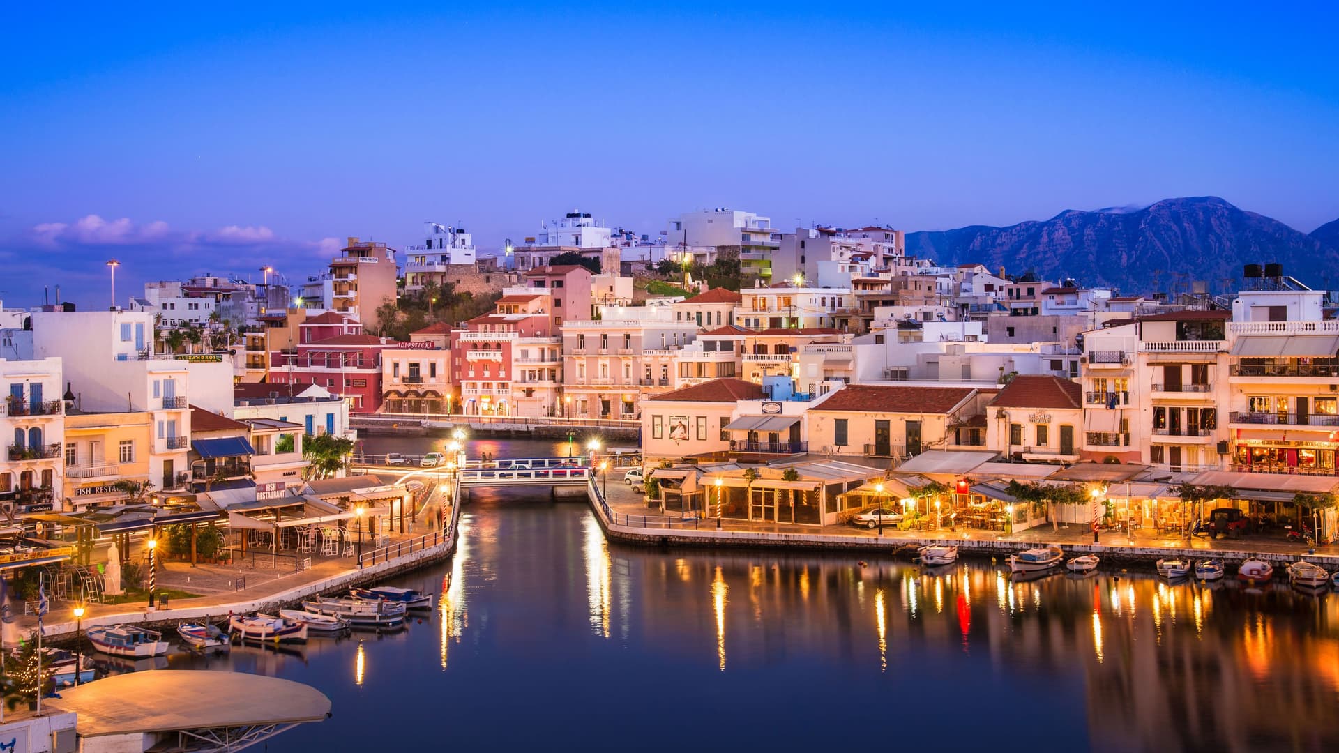 A stunning dusk shot of Agios Nikolaos, Crete, with colorful buildings and restaurants lining the tranquil Voulismeni Lake, their lights reflecting on the water.