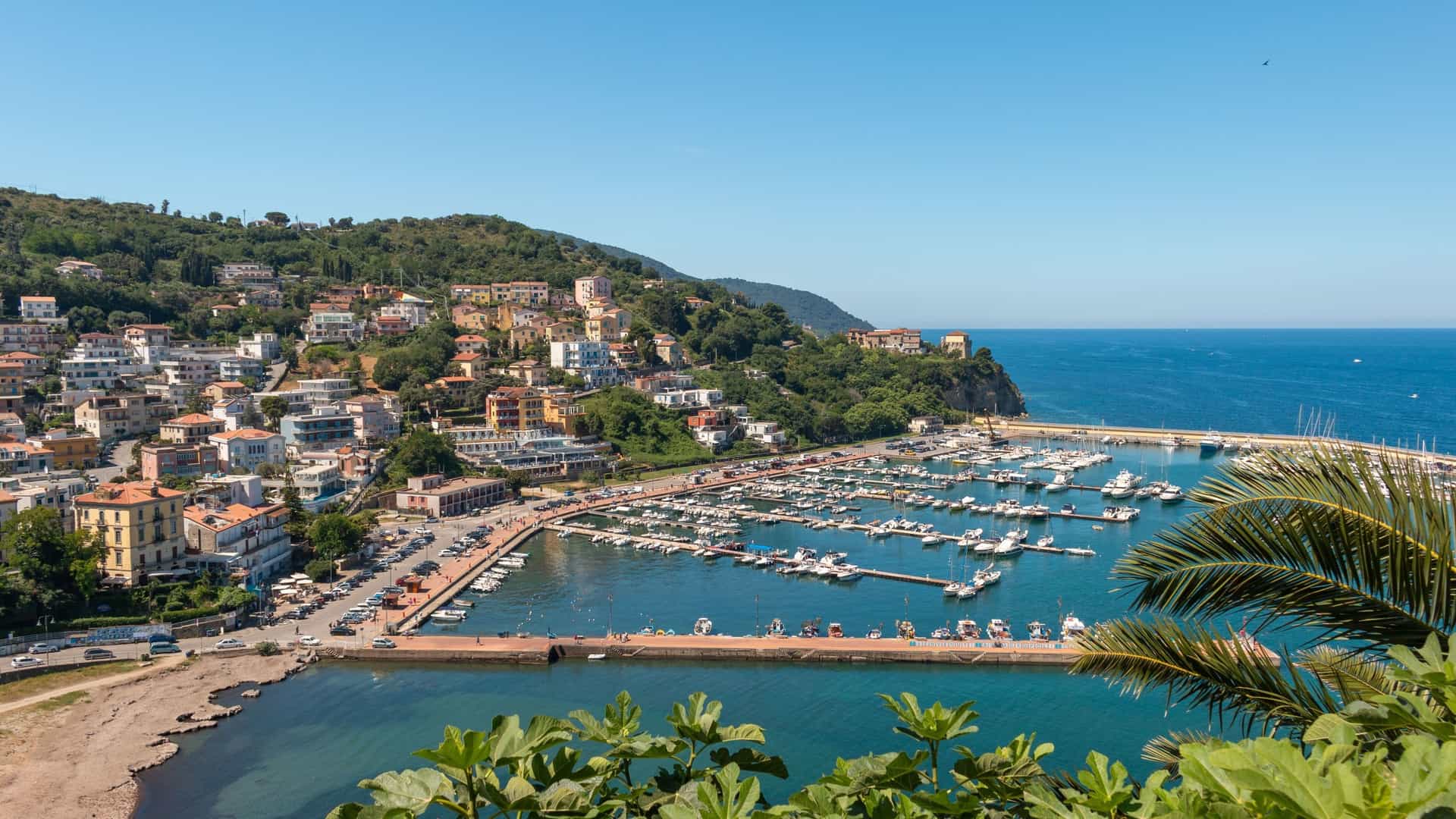 A high-angle view of the bustling Agropoli marina in Italy, with numerous boats moored in the harbor, surrounded by a picturesque town built into a green hillside.