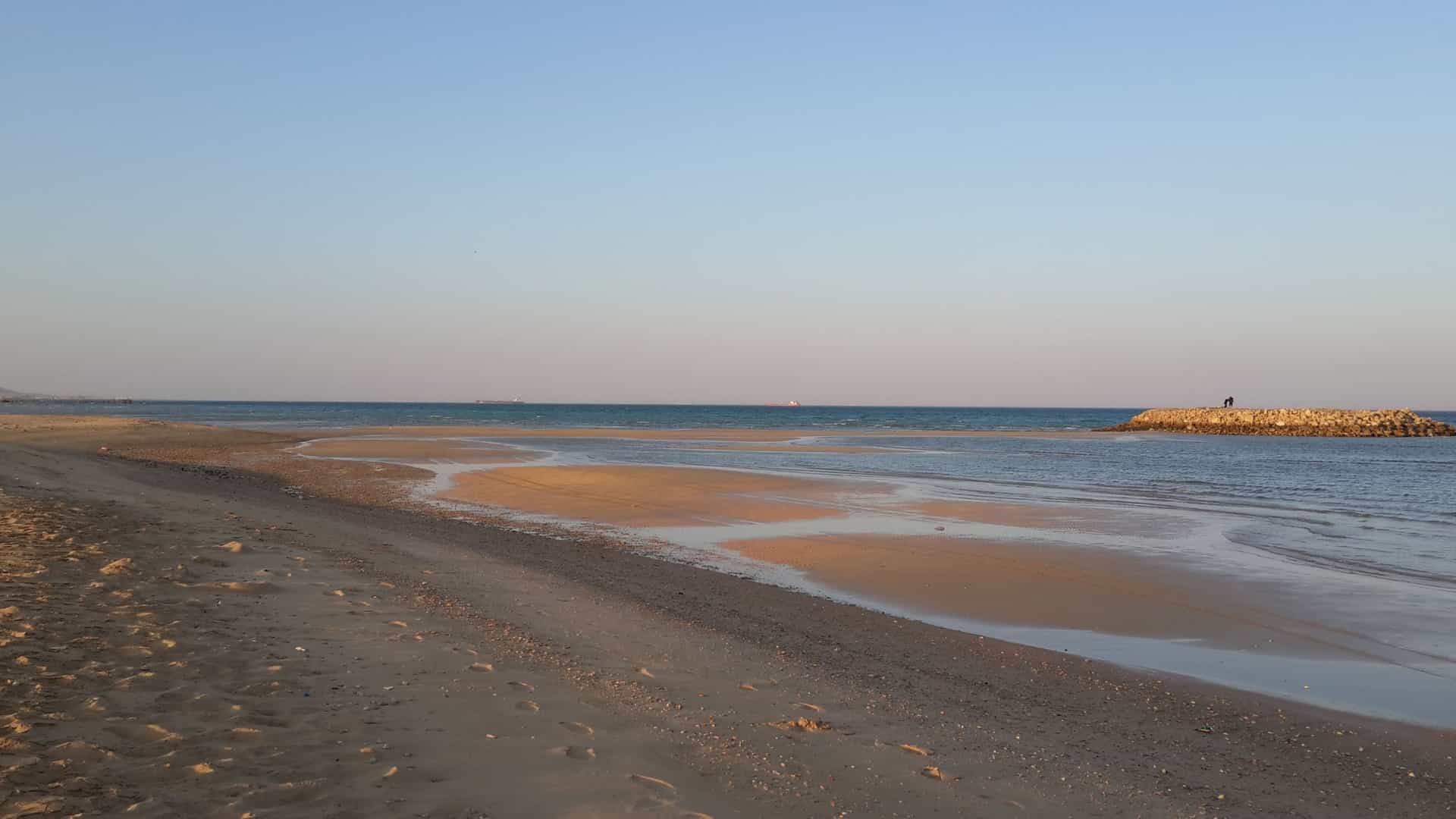 A calm beach scene in Ain Sukhna, Egypt, showing the Red Sea at low tide with a vast expanse of sandy shoreline and a small pier in the distance.