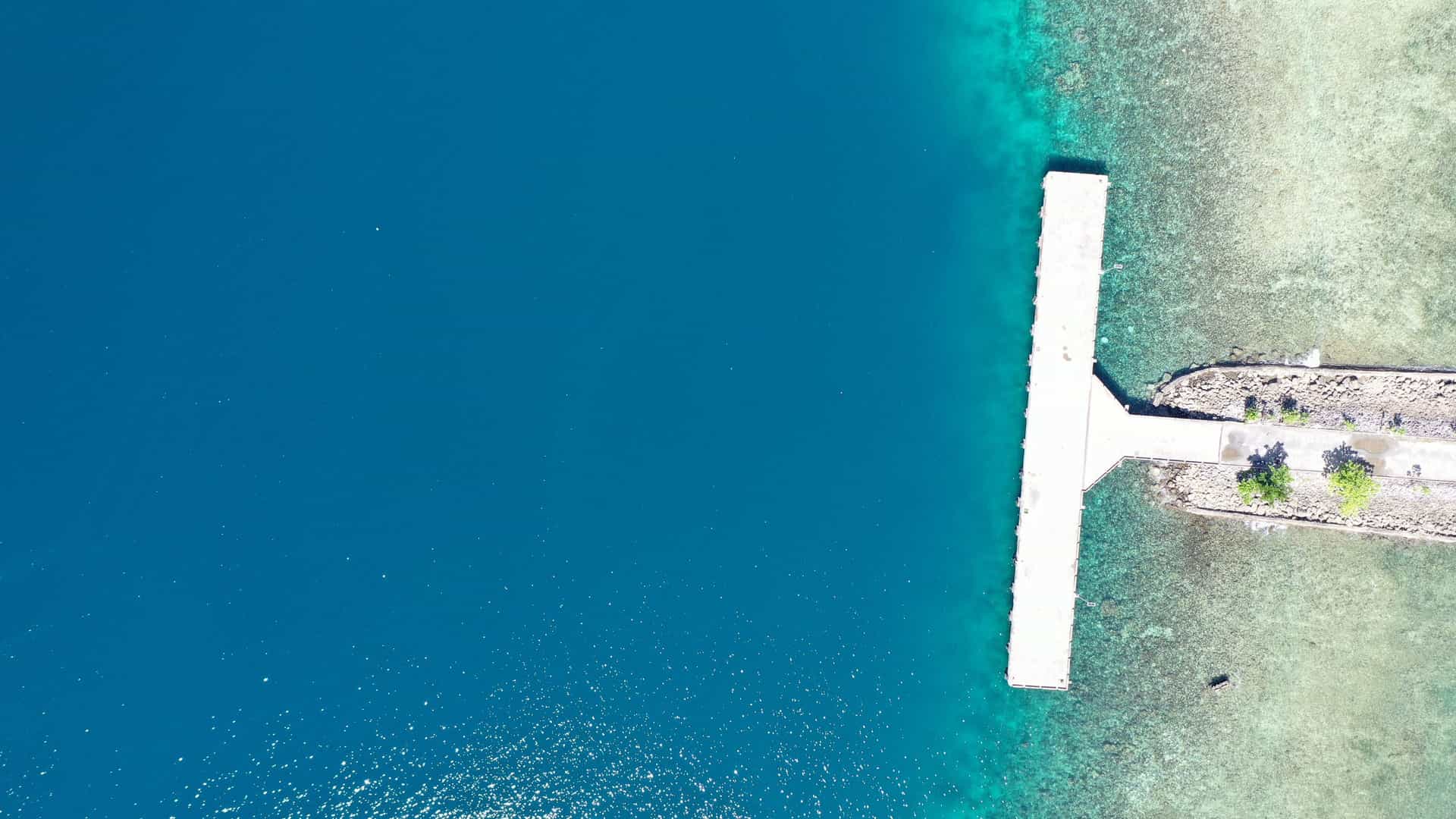 An aerial, top-down view of a concrete pier in Aisandami, Indonesia. The pier extends from a rocky shoreline into the ocean, which transitions from a shallow, clear turquoise over a reef to a deep, dark blue.