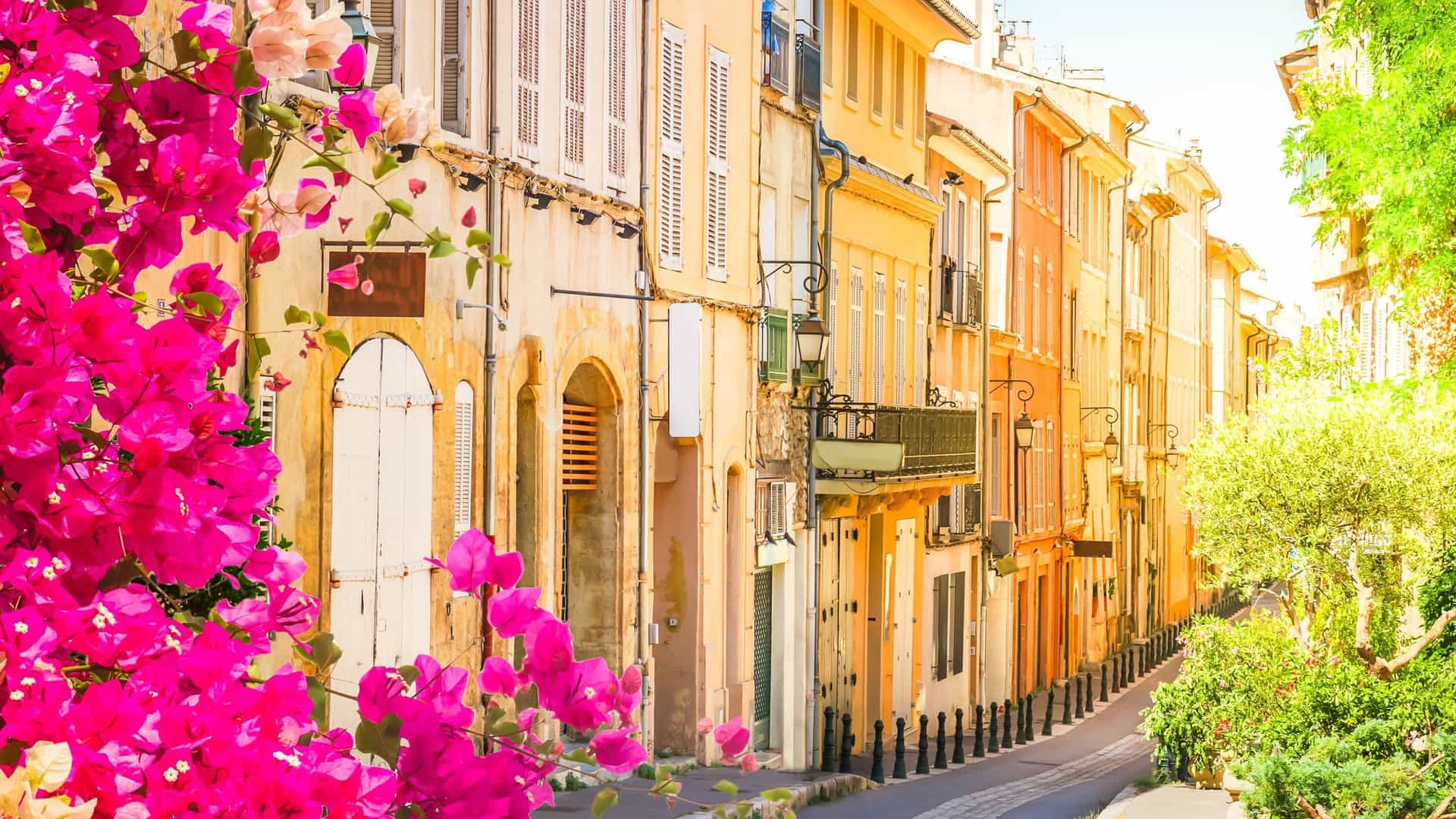 A picturesque street scene in Aix-en-Provence, France, with a row of historic pastel-colored buildings and vibrant pink bougainvillea flowers in the foreground.
