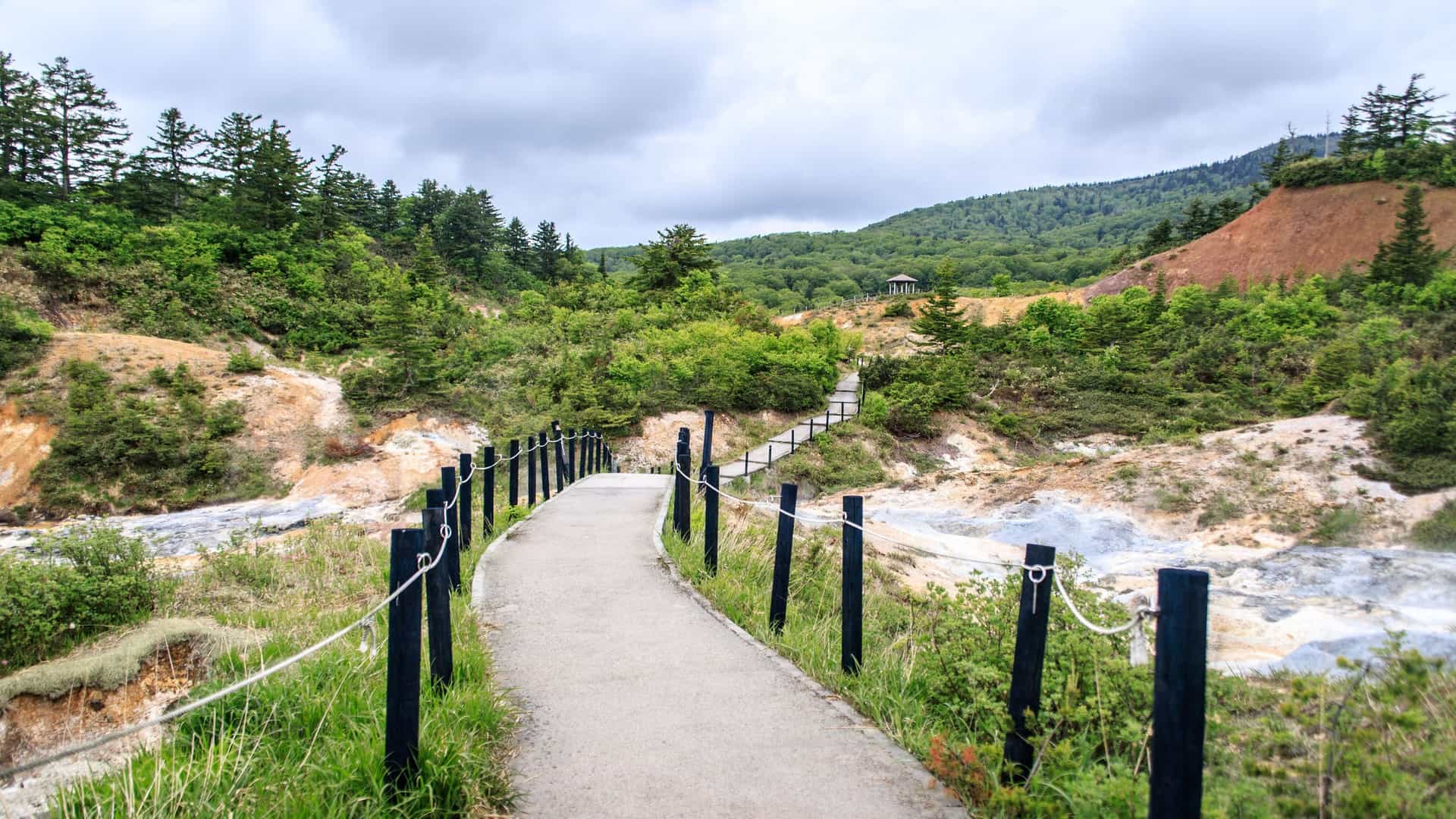 A winding elevated walkway with a rope fence leads through the volcanic landscape of Akita, Japan, with green trees and a small gazebo visible in the distance.